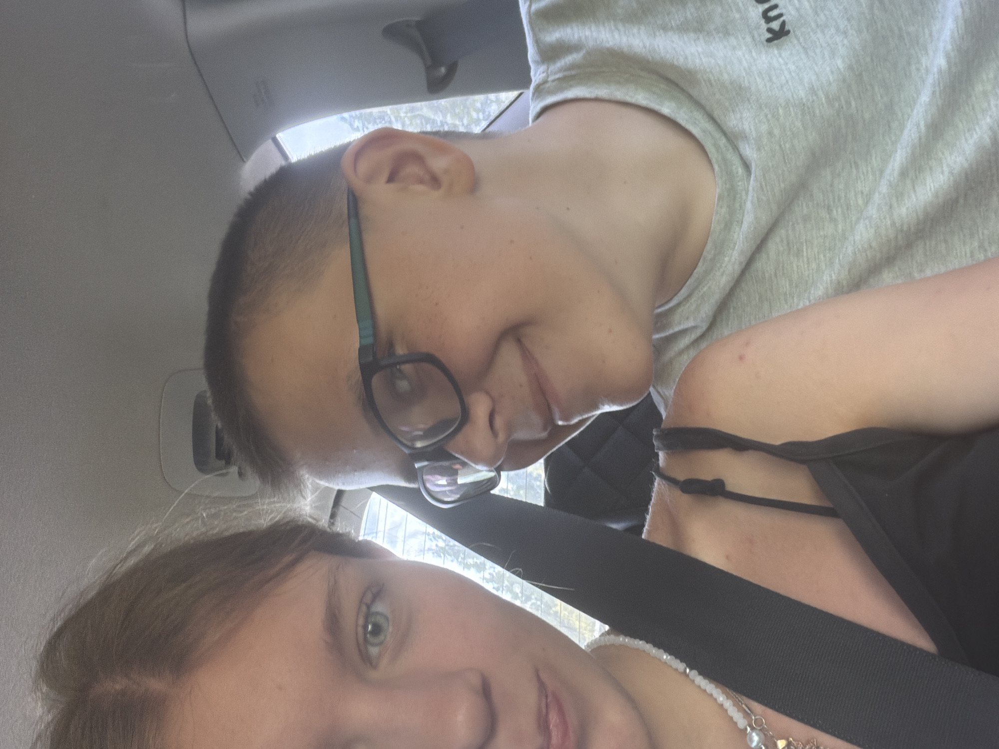 Two individuals, a female on the left and a male on the right, are positioned in the front seats of a vehicle, likely a car, facing the camera. The female, partially visible on the left, has light brown hair and light blue eyes, and is wearing a black camisole and a beaded necklace. The male, to the right, has short brown hair, wears glasses with green frames, and is dressed in a grey t-shirt with black lettering on the right side. Both individuals appear to be smiling. The background is blurred, but suggests an indoor setting within a vehicle, with glimpses of the outdoors visible through the car windows. The image quality is moderate, with natural lighting. The provided location, Novaya Zhizn', Russia, is not visually discernible from the image itself.