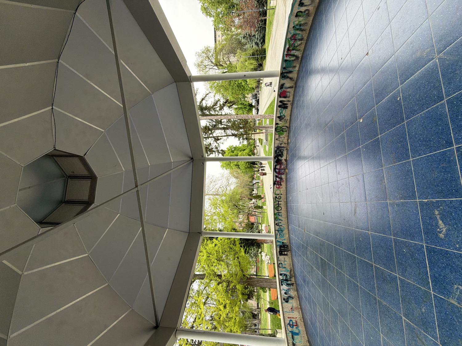 The image, correctly oriented, depicts the interior of a large, circular, open-air pavilion or gazebo, looking out into a public park. The ceiling of the structure is a complex, grey, paneled octagonal design, featuring a central vent. Below, the floor is covered with numerous small, square blue tiles, forming a wide, circular area, suggesting a stage or communal space. This tiled floor is bordered by a low, curved concrete wall or railing that is heavily covered with multi-colored graffiti, indicating a history of public use and expression.
Beyond the pavilion's structure, a vibrant green park is visible under bright daylight. Lush trees with full foliage, grassy areas, and several park benches are scattered throughout. In the mid-ground, some white, possibly statue-like figures (perhaps swans or ducks) can be seen on the grass. Further in the background, residential or public buildings are faintly visible, along with what appears to be a road or pathway and parked cars. The weather appears clear and bright, consistent with a daytime scene, likely in spring or summer. No people are prominently visible within the frame. The overall atmosphere suggests a well-used urban park space, possibly in Pleven, Bulgaria, where the pavilion stands as a central feature. The graffiti adds a raw, urban element to the otherwise natural park setting.