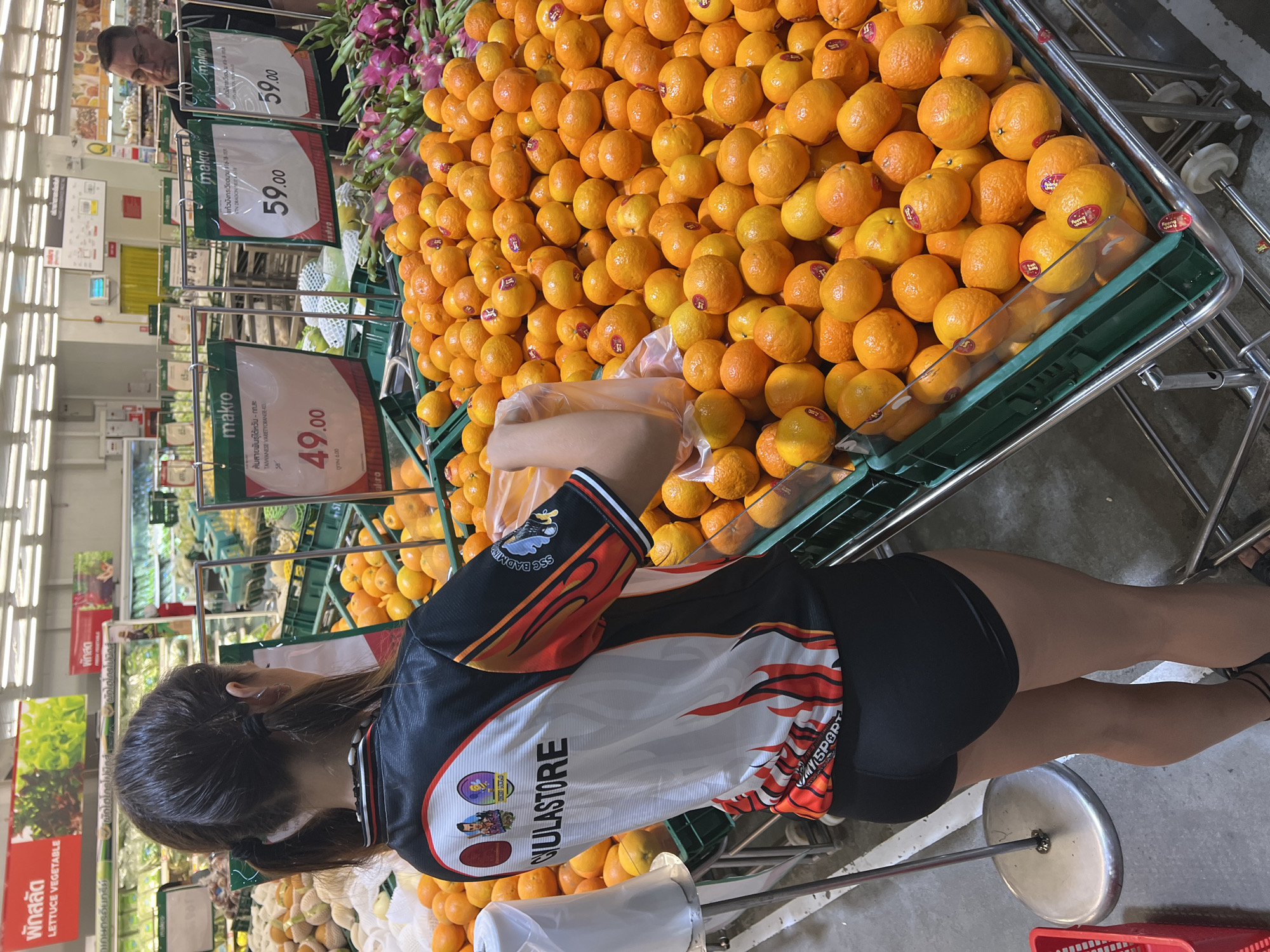 The image captures an indoor scene at Makro in Baan Bo Phut, Thailand. A woman wearing a black shirt and shorts is picking oranges from a large display cart filled with them. The cart is green and has wheels. Above the cart are price signs displaying "49.00" and "59.00" in Thai script and English. A man is standing near the cart in the background. Also visible in the background are signs indicating "LETTUCE VEGETABLE" in English and Thai. The floor is gray concrete.