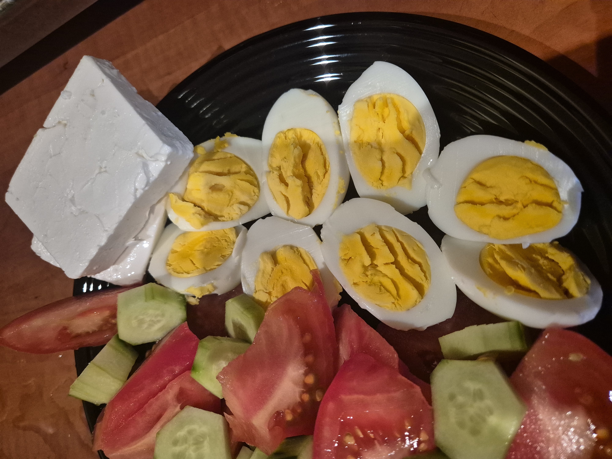 A close-up, overhead shot reveals a plate filled with food, likely prepared for a meal. The central subjects are various ingredients neatly arranged on a black, ridged plate.
On the upper right and center of the plate, there are several halves of hard-boiled eggs, showcasing their bright yellow, fully cooked yolks and firm white albumen. To the left, a substantial block of white cheese, possibly feta or a similar brined cheese, sits prominently, displaying its crumbly texture. Below the eggs and cheese, occupying the lower portion of the plate, is a mix of freshly chopped vegetables: vibrant red tomato pieces and green cucumber slices.
The plate itself is dark black with distinct concentric ridges, adding a subtle texture to the dinnerware. The entire arrangement rests on a warm, brown wooden surface, which serves as the background. The lighting appears to be even, highlighting the colors and textures of the food. No activity is explicitly depicted beyond the presentation of the meal. There are no people visible, and no text is discernible in the image. The scene suggests a wholesome and fresh, perhaps Mediterranean-inspired, breakfast or light meal.