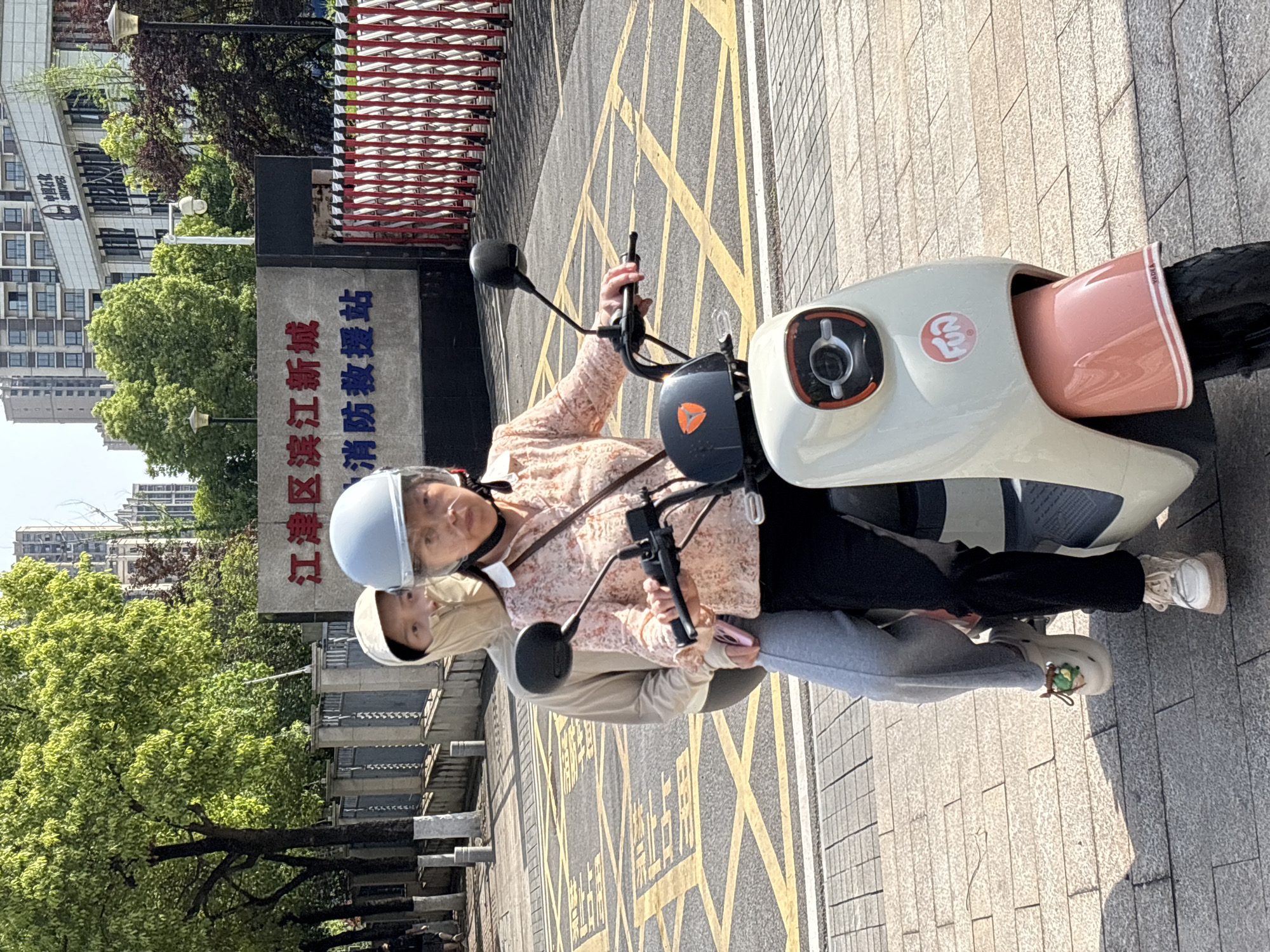 Two individuals are pictured on a white and pink electric scooter, seemingly paused on a paved urban street. An older woman, wearing a light-colored helmet and a floral patterned top, is in the driver's seat, holding the handlebars. Behind her, a younger person, dressed in a beige hooded top, is seated as a passenger. The scooter features a "Yadea" logo on its front and a red circular logo, possibly a brand identifier.
The scene is set outdoors under bright daylight, indicated by clear shadows on the ground. The road surface has prominent yellow crosshatch markings, typically indicating a no-stop zone or a specific road instruction. Adjacent to the road is a sidewalk, and in the background, there are green trees and several multi-story buildings.
A large grey sign to the left of the scooter bears Chinese characters, reading "区江江新城 消防救援站" (Jiangjin District New City Fire and Rescue Station) in red and blue text. Another building in the background has "中国民生" (China Minsheng) visible on its facade. The image captures an everyday moment of transportation in what appears to be Jiangjin, China.