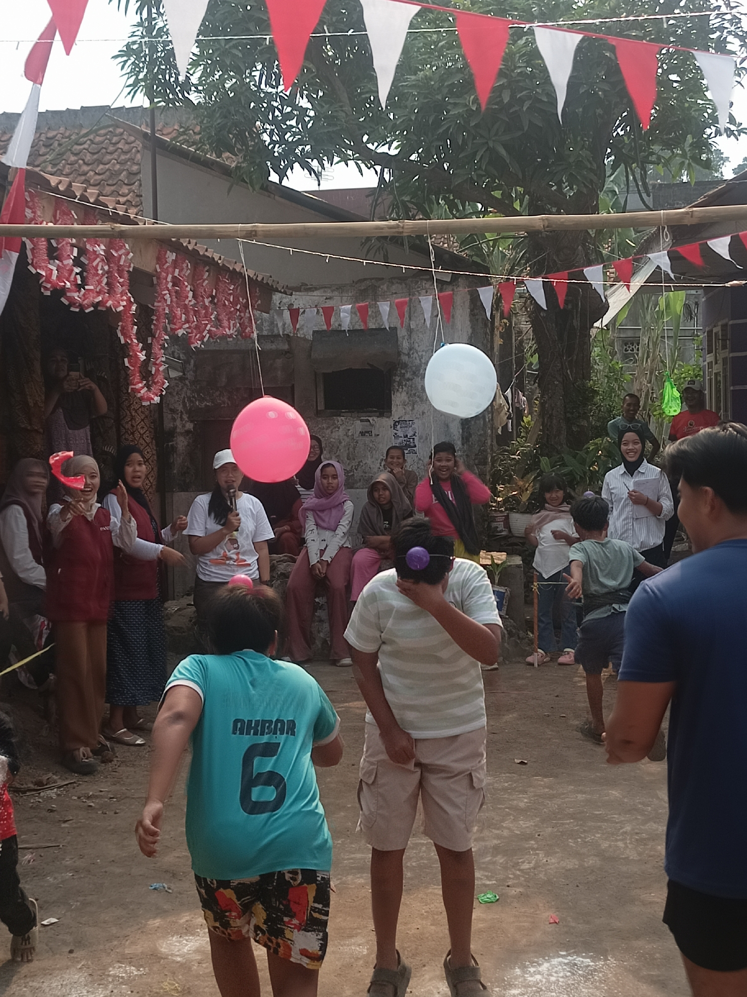 The image depicts an outdoor gathering, likely a community event or festival, in Tasikmalaya, Indonesia. The scene is decorated with red and white triangular flags and garlands, commonly used during Indonesian Independence Day celebrations. Several people are present, including men, women, and children.
In the foreground, a young boy wearing a teal t-shirt with the name "AKBAR" and the number "6" on the back is running. He is wearing patterned shorts and sandals. To his right, a person in a striped t-shirt and beige shorts is facing away from the camera, possibly participating in an activity.
In the middle ground, a woman wearing a white shirt and a cap is holding a microphone and appears to be speaking. She is standing near a large pink balloon that is suspended in the air. Other individuals, including children and adults, are observing the event. Some participants are seen with what appear to be small objects attached to their heads or faces, suggesting a game or competition.
The background shows a building with a tiled roof and some trees. The ground is dusty and uneven. The overall atmosphere suggests a festive and participatory event.