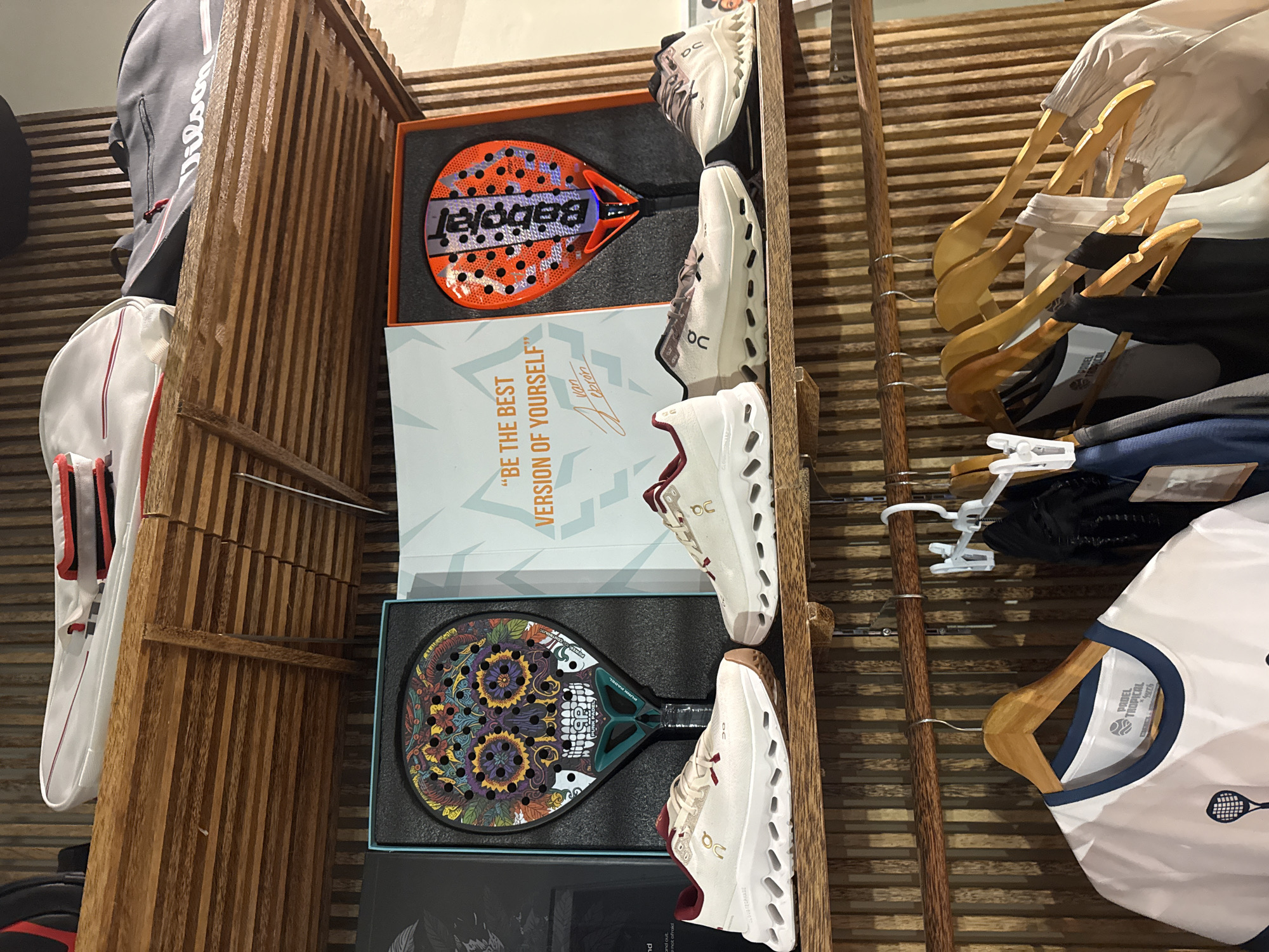 This media file showcases a display of padel tennis equipment and apparel. The primary subjects are padel rackets, shoes, and clothing, presented in a retail or exhibition setting. The scene is composed of several items, including two padel rackets in their boxes, one with an orange and black design featuring the "Babolat" logo, and another with a colorful skull and floral motif. Several pairs of white athletic shoes with dark accents are also visible, stacked vertically.
Adjacent to the equipment, there is a white box with text that reads, "BE THE BEST VERSION OF YOURSELF" by "Juan Lebron". A grey and white Wilson brand bag is positioned on the left side of the frame. Hanging wooden and wire hangers with various pieces of clothing, including a white t-shirt with "PADEL TROPICAL" printed on the label, are seen on the right.
The setting appears to be indoors, with a wooden slatted wall providing a textured background. The lighting suggests it could be daytime, with no specific indication of outdoor weather conditions. The overall presentation is neat and organized, characteristic of a sports store or a dedicated display area. No people are visible in the frame. The visible text provides brand names and motivational quotes related to the sport.