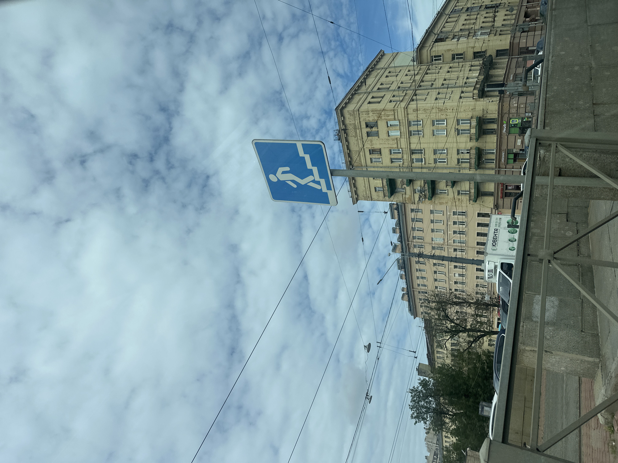 A street scene in Saint Petersburg, Russia, captures a cloudy sky with scattered white clouds and blue patches. Overhead, a network of black electrical wires crisscrosses the frame. In the foreground, a metal railing with a grey stone barrier runs horizontally. A blue square sign with a white silhouette of a person walking up stairs is mounted on a pole. Behind the sign, a tall, light yellow apartment building with multiple floors and windows dominates the background, displaying signs of age and weathering. To the left of this building, another, slightly shorter apartment building of a similar architectural style is visible. A white van with Russian text and a logo is parked on the street, along with several other cars. Green trees are present in the midground.