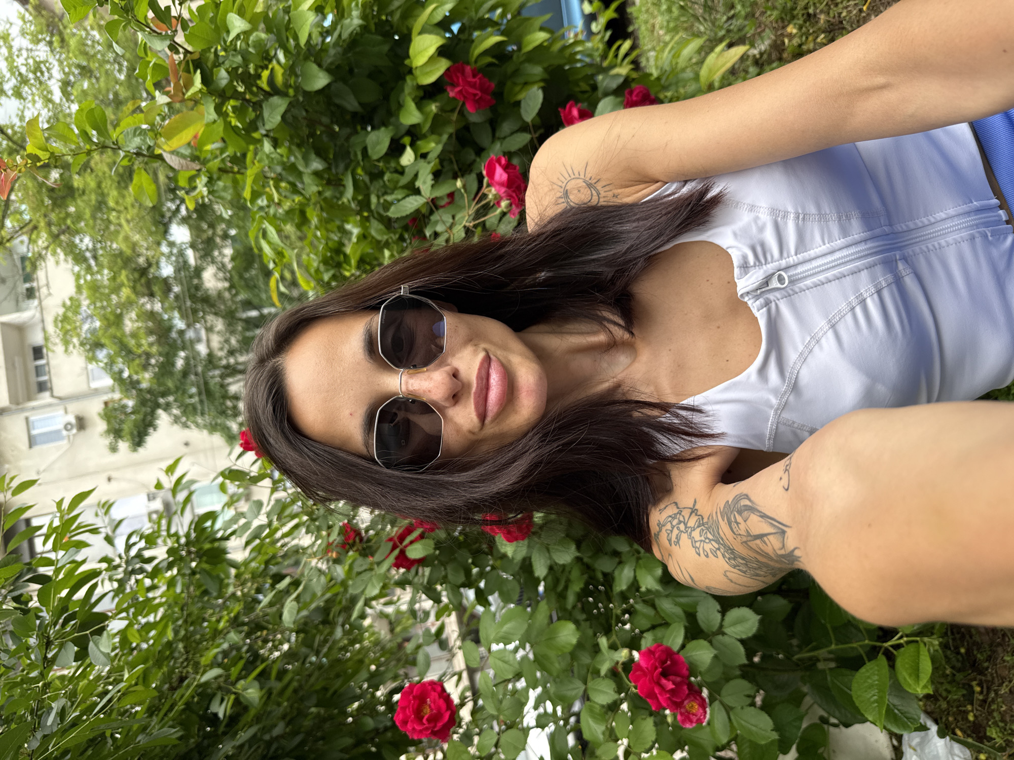 The image shows a woman with long dark hair wearing sunglasses and a white top, posing in front of a rose bush with red flowers. The location appears to be in Pleven, Bulgaria, based on the visible building in the background. The woman has tattoos on her arms and is facing the camera. The background includes green foliage and glimpses of urban buildings.