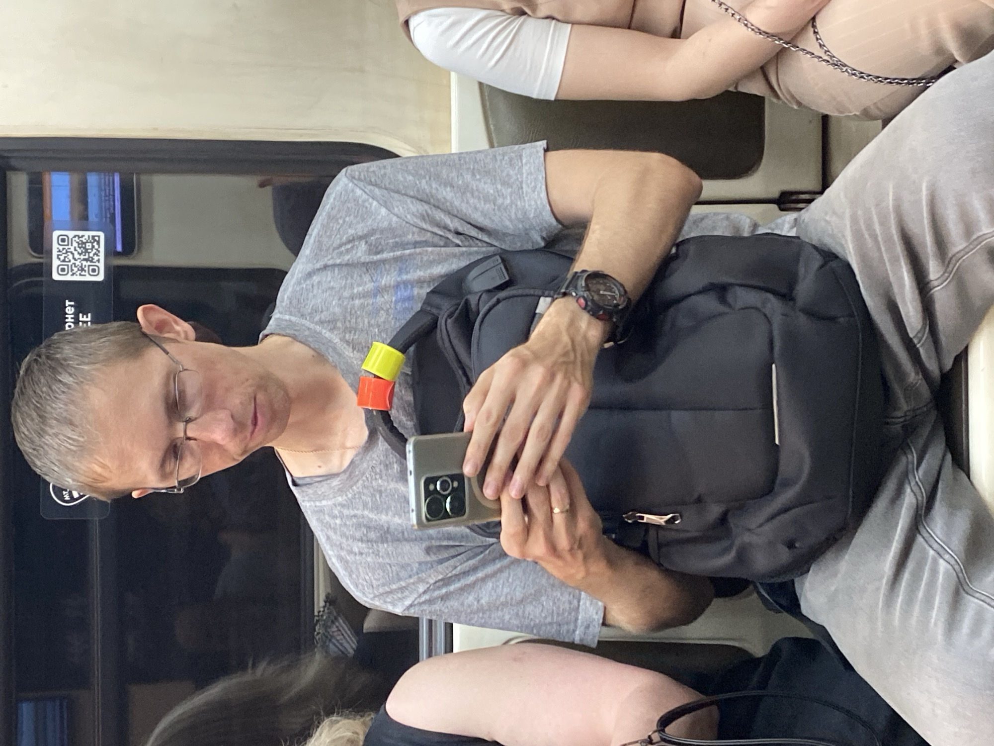 The image captures a man with short hair and glasses, wearing a grey t-shirt and grey pants, sitting on a train. He is holding a smartphone in his hands and looking down at it. A black backpack with a yellow and red strap is positioned behind him. To his left, a woman with blonde hair is partially visible, wearing a black top and holding a black handbag. To his right, a woman in a beige top and matching pants is also visible, her arm resting on her leg. The background suggests an indoor public transportation setting, possibly a metro train car, with a dark window reflecting the interior and a sign with Cyrillic text and a QR code. The lighting is moderate, and the overall scene is devoid of any discernible interaction between the individuals.