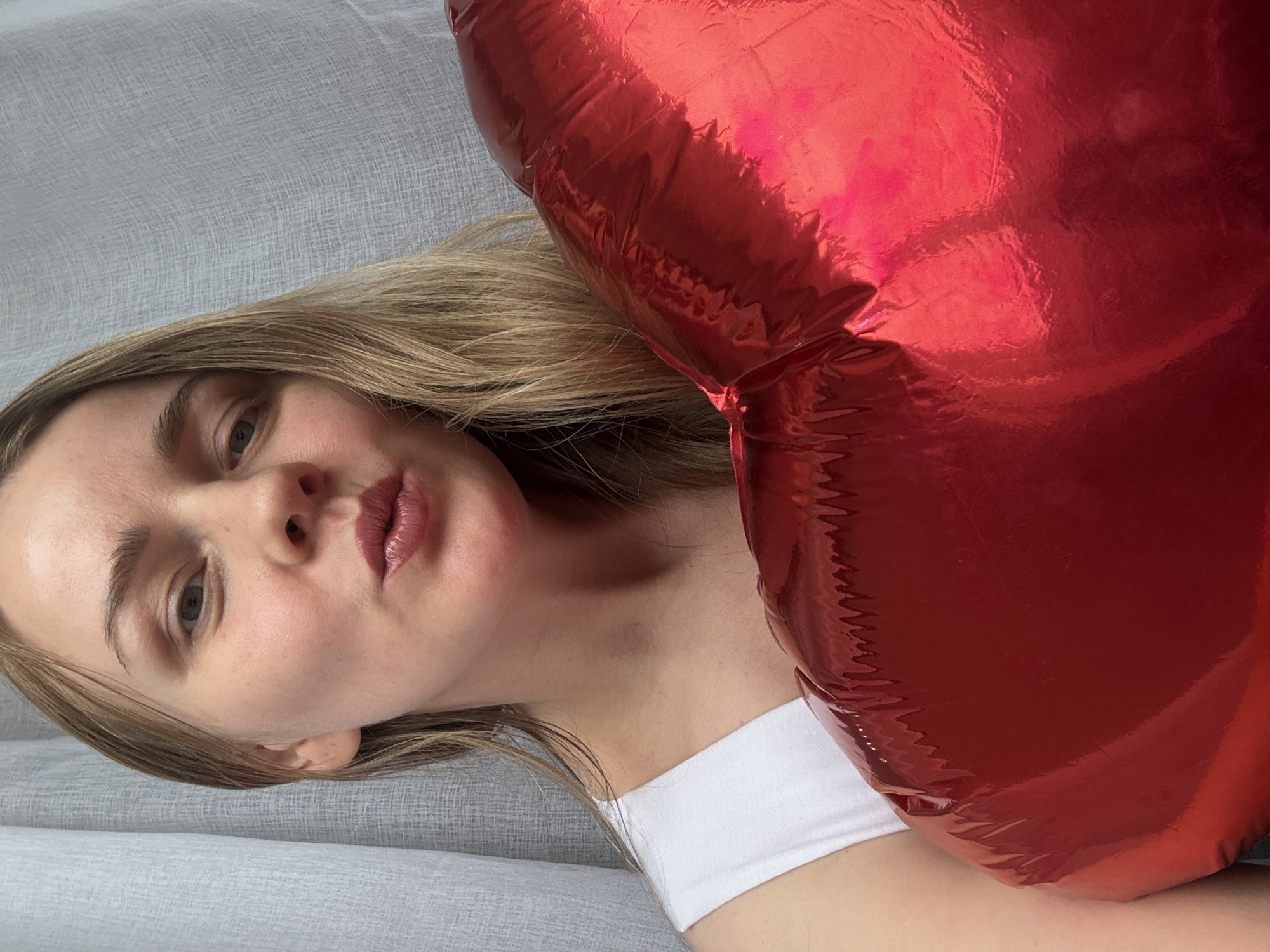 A woman with blonde hair and fair skin is posing with a large, red, heart-shaped foil balloon. She is wearing a white top. The background appears to be a light gray fabric surface.