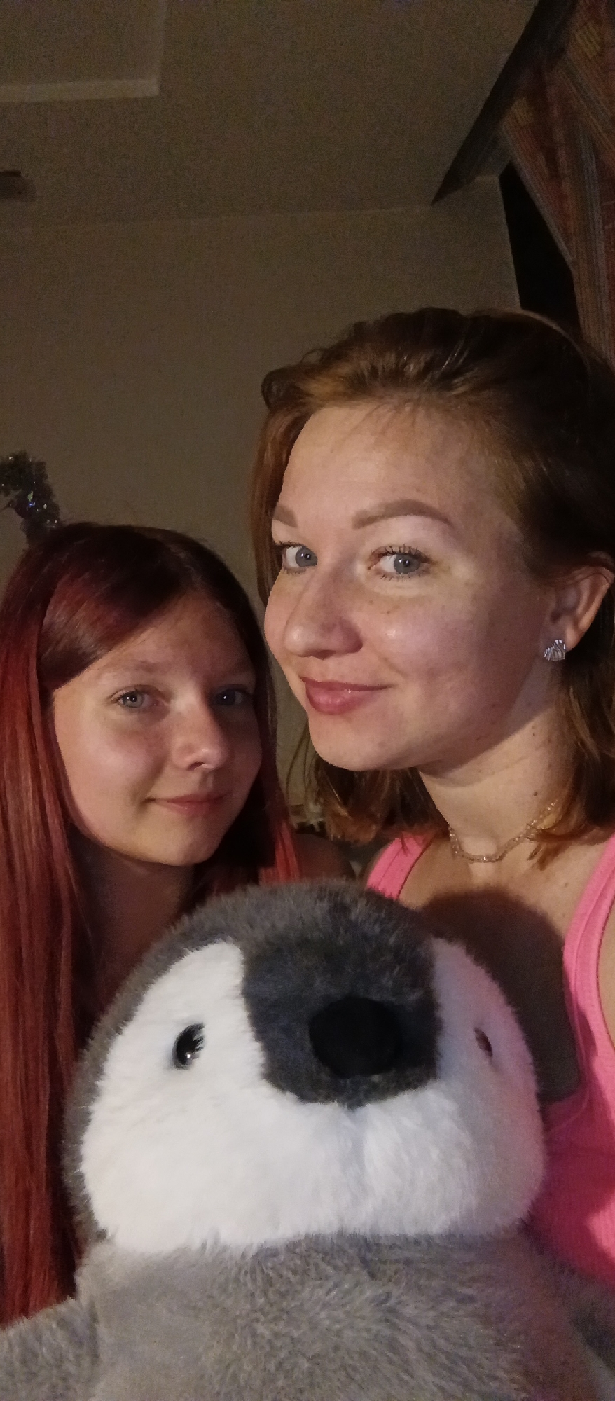 Two women, one with red hair and the other with reddish-brown hair, are posing for a photograph while holding a stuffed penguin. The woman with red hair is on the left, and the woman with reddish-brown hair is on the right. The woman on the right is wearing a pink tank top and a delicate necklace, and she has small, shell-shaped earrings. The background is nondescript, with a hint of a patterned curtain visible on the right. The location is Unknown, Unknown.