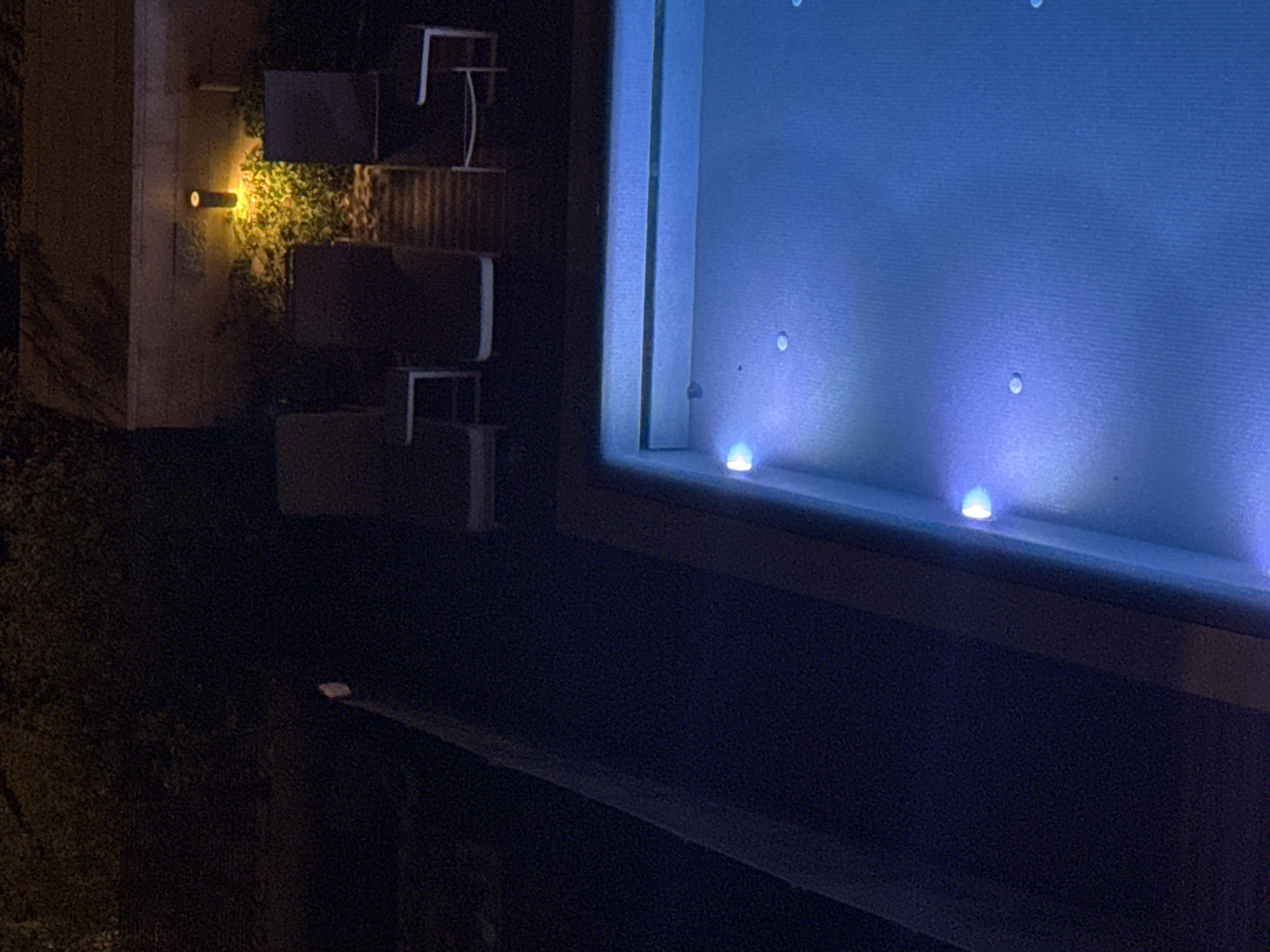 The image appears to be a nighttime shot, likely taken from within a building, overlooking an outdoor patio or garden area. The location is unknown as to whether it is in Tel Aviv, Israel.
To the left of the frame, there's a textured, vertical wall with what appears to be a landscape light fixture casting a warm yellow glow. Adjacent to this wall is what seems to be a patio area. It features dark outdoor furniture, including chairs and possibly a table. There is foliage visible, receiving light from the landscape light.
The foreground is dominated by the interior of the building. There's a window sill with blue lights placed along the bottom edge, emitting a strong light. The wall is dark and appears to be painted a deep blue color. The window itself is large and reflects the blue light, creating an illuminated area. The focus is more on the light and shadow play rather than the objects themselves.