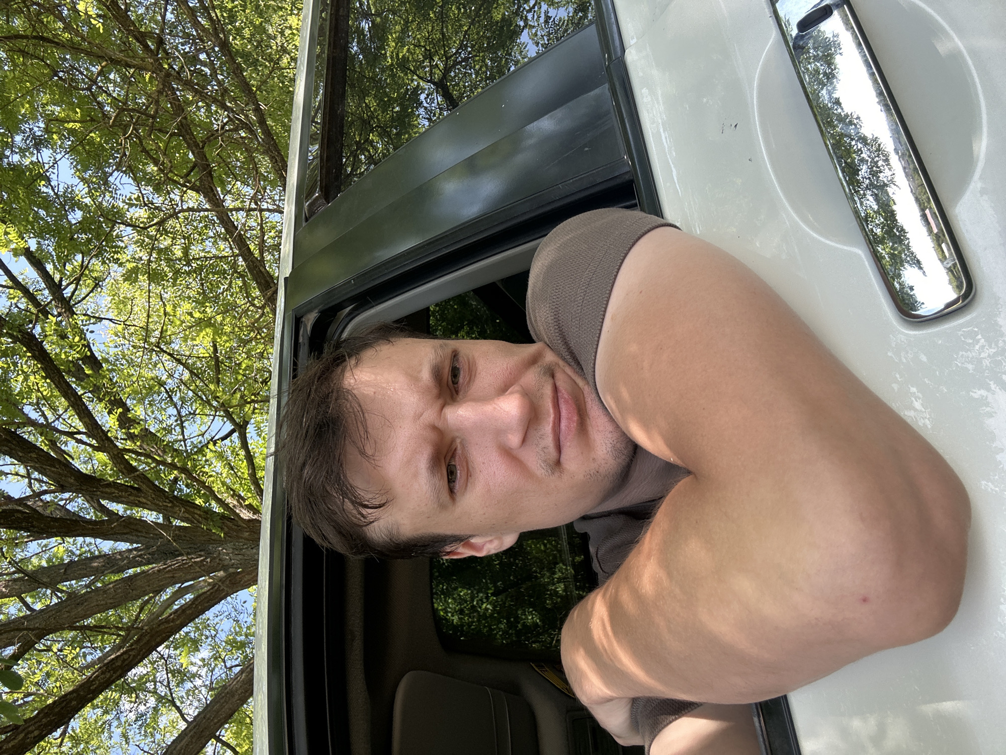 A man is leaning out of a white car window, his left arm resting on the exterior of the car. He has short, dark brown hair and is looking directly at the camera with a pensive expression. The car is parked outdoors, with a large tree and green foliage visible in the background, and the sky above. The reflection of trees and buildings can be seen in the car's window and chrome door handle. The scene is likely in Makiv, Ukraine, given the presence of trees and what appears to be a rural or suburban setting.
