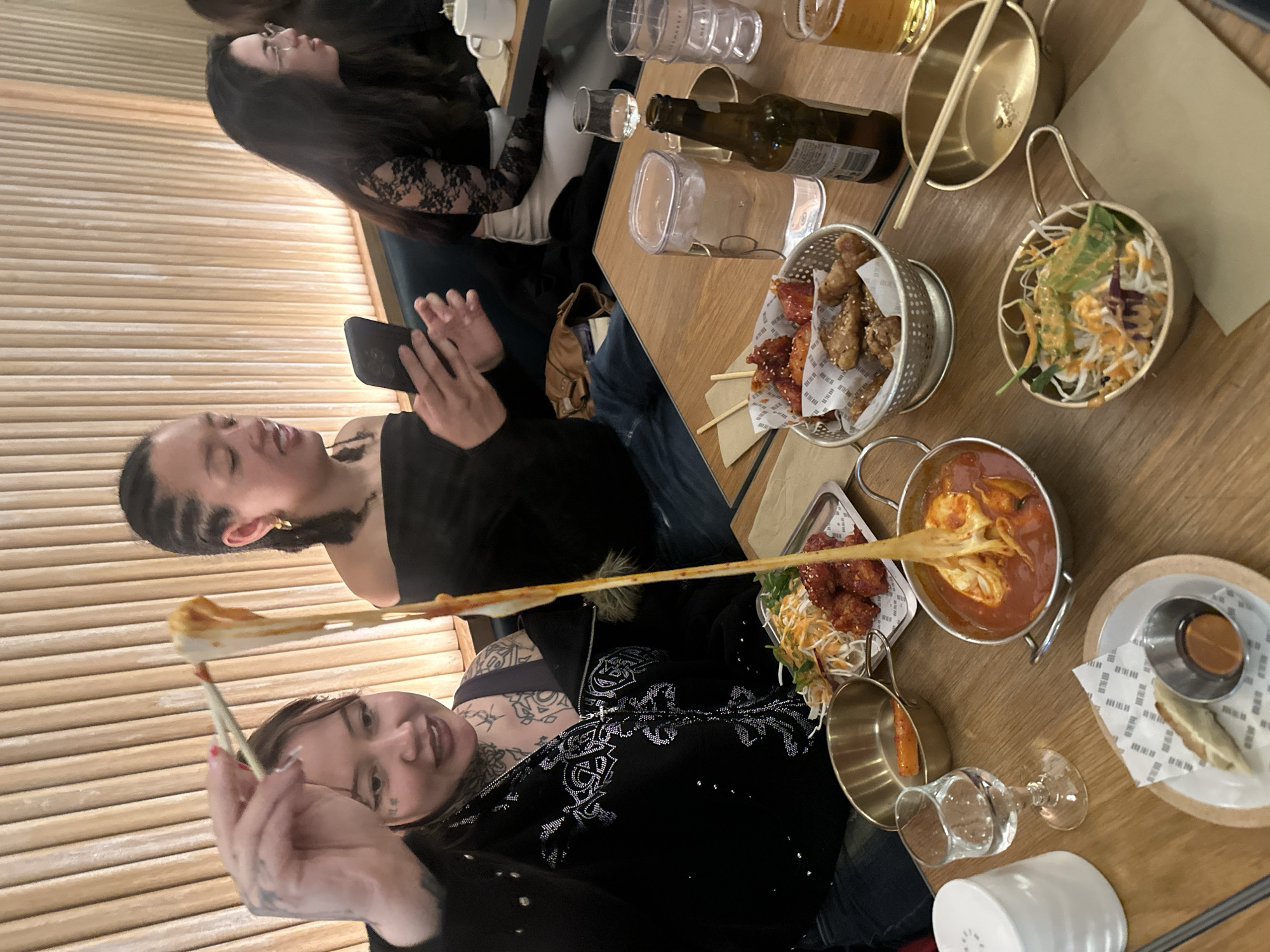 The image depicts an indoor dining scene at a restaurant, likely a Korean establishment in London, with three individuals enjoying a meal together. The perspective is an overhead, tilted shot of the table and diners.
In the foreground, a person with dark hair and tattoos on their arm and chest is actively engaged in eating, using chopsticks to pull a long, stringy strand of melted cheese from a pot of red stew, consistent with Tteokbokki. They are wearing a dark, embellished top and show a faint smile of satisfaction. To their left, another individual with dark, braided hair is holding a black smartphone, seemingly engrossed in it. Further back, partially visible, is a third person with long dark hair, wearing a dark top with lace sleeves.
The wooden table is covered with an assortment of dishes and drinks. Prominently featured is the pot of Tteokbokki with the stretching cheese. Alongside it, a metal basket holds pieces of Korean fried chicken, some coated in a spicy red sauce and others sprinkled with sesame seeds. There's a small, gold-colored bowl containing a fresh salad with dressing, and another small bowl with a single rice cake. Other items include small metal ramekins with sauces, a plate with fried items and a dipping sauce (on which the text "THE NEW" is visible), and several glasses, including one with a beverage resembling beer, and a dark glass bottle. The background features warm, horizontally paneled wooden walls, suggesting a modern and casual dining environment. The overall mood is relaxed, centered around shared food and conversation.