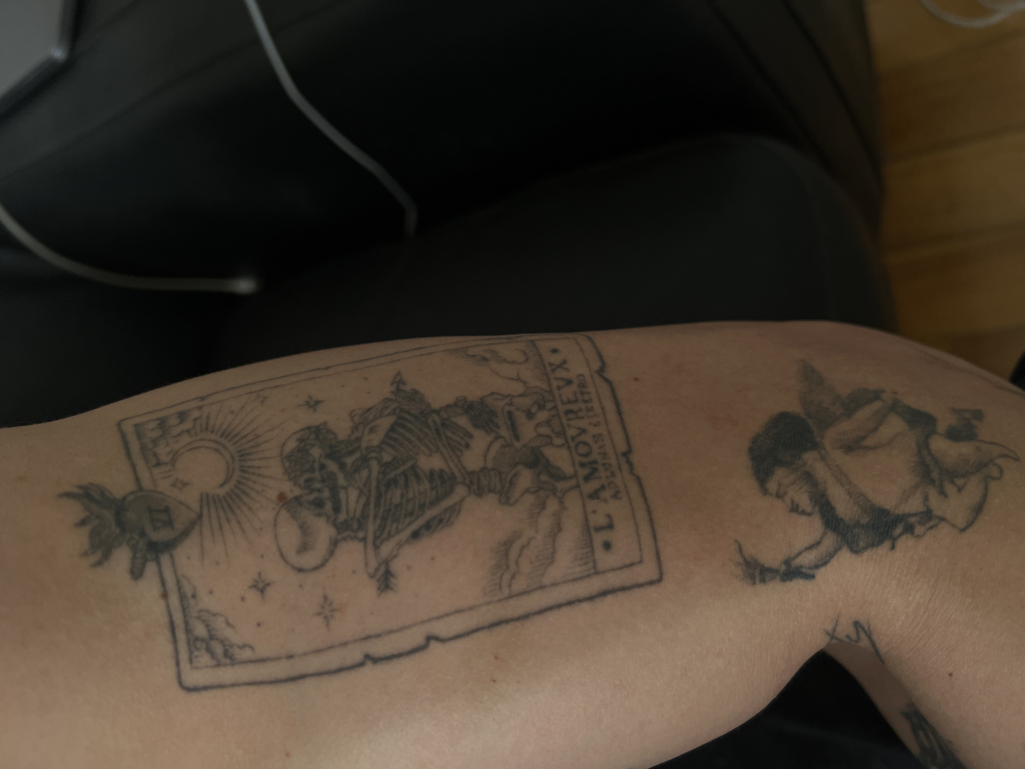 A close-up shot captures a person's arm, heavily adorned with intricate black ink tattoos, set against a blurred, dark background that includes faint glimpses of white wires and a light wooden surface. The arm itself appears to be resting comfortably.
The most prominent tattoo is a large, rectangular design resembling an antique print or tarot card. Within its detailed frame, two skeletal figures are depicted in an embrace or dance, under a radiant sun and a crescent moon, surrounded by small stars. To the left of this main frame, a smaller, stylized winged heart or fruit-like object bears the inscribed letters "EI" or "GI". The main tattoo also features distinct text: "L'AMOVREVX" is clearly visible, likely an archaic spelling of "L'AMOREUX" (The Lover), a known Tarot card. Below it, the words "ADORNAM & RETRO" are inscribed vertically.
Further along the arm, to the right of the larger tattoo, a second, equally detailed tattoo portrays an angel or cherubic figure with dark hair, seemingly in motion or suspended in air. A smaller, less clear tattoo is partially visible further down the arm. The overall impression is one of deeply personal artistry. This scene is set in Mexico City, Mexico.