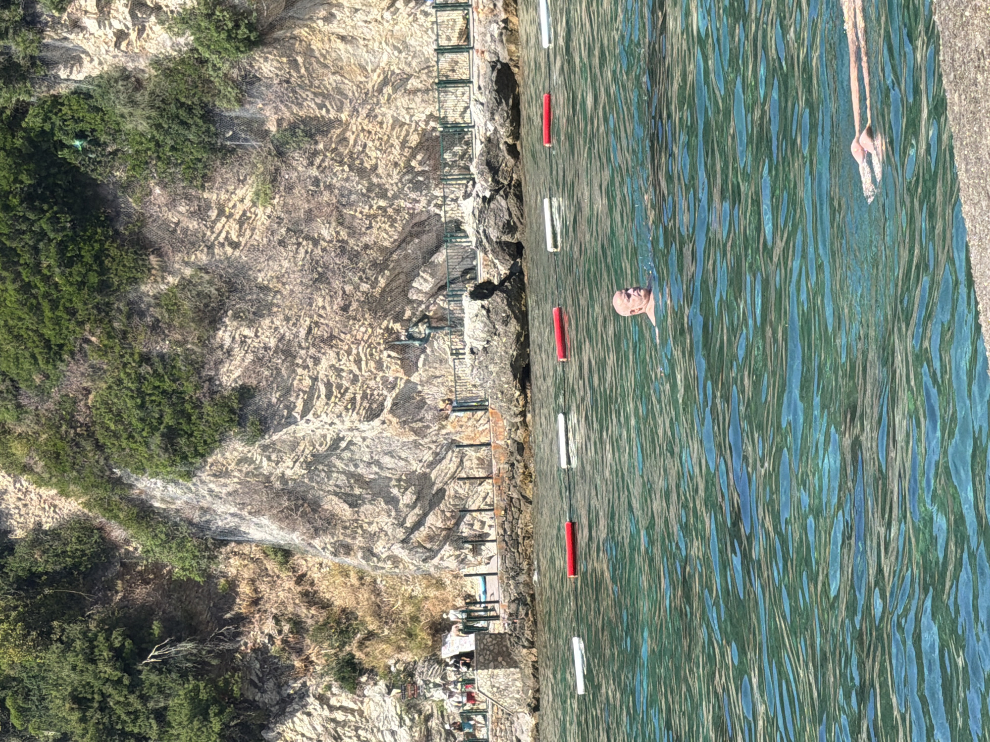 This is a photograph taken at Mogren Beach in Budva, Montenegro. The image captures a section of the Adriatic Sea with clear turquoise waters. In the foreground, a man with a shaved head is swimming, his head and shoulders visible above the water. Further to the right and closer to the bottom of the frame, a pair of legs are visible just below the water's surface, indicating another swimmer. A rope with alternating red and white buoys demarcates a swimming area in the sea. In the background, a rocky cliff face covered in dense green vegetation rises steeply. Along the base of the cliff, a paved walkway or promenade is visible, bordered by a metal fence. Several people are standing or walking along this promenade, some appearing to be near a small stone structure. The overall scene depicts a sunny day at a coastal resort.