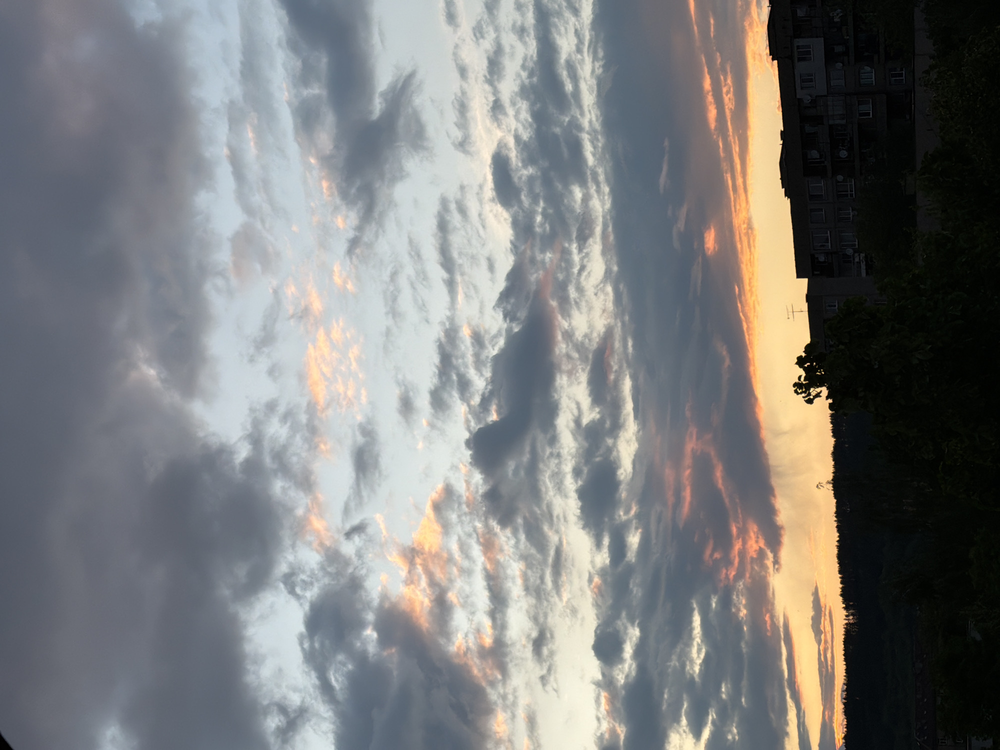 The image captures a sky at sunset in an unknown city. The sky is filled with clouds, some dark gray and others illuminated with orange and yellow hues from the setting sun. A building and trees silhouette the skyline on the right.