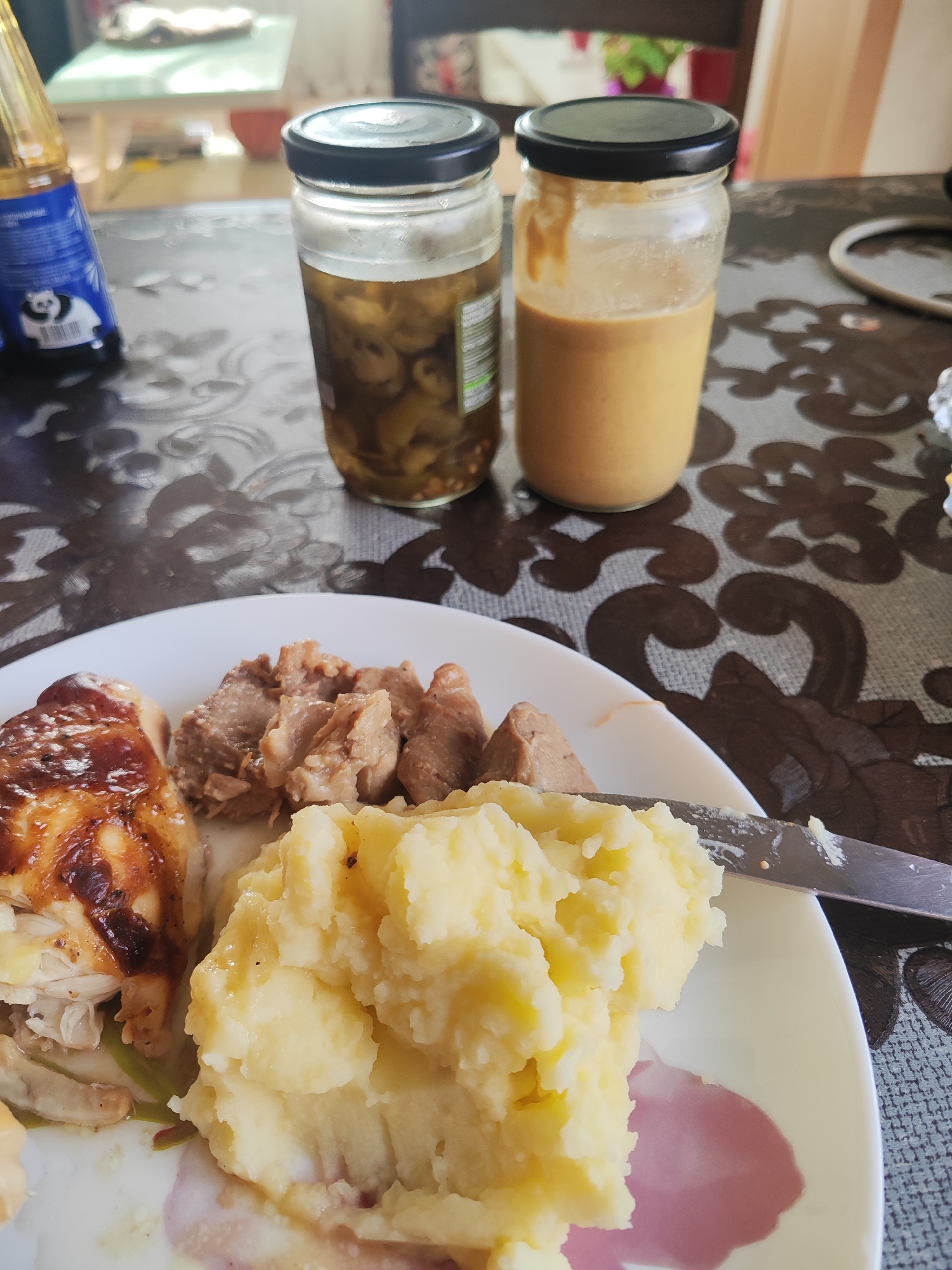 A close-up, eye-level shot shows a plate of food with two glass jars and a bottle on a patterned tablecloth. The plate contains mashed potatoes, pieces of cooked meat, and what appears to be roasted chicken. The two glass jars have black lids, and one contains sliced pickles while the other holds a thick, beige sauce. A bottle with a blue label featuring a panda illustration stands to the left. The scene is indoors, with a table, chairs, and a window visible in the background. The lighting suggests it's daytime. No individuals are present in the image. This scene was captured in the city of Gvardeysk, Russia.