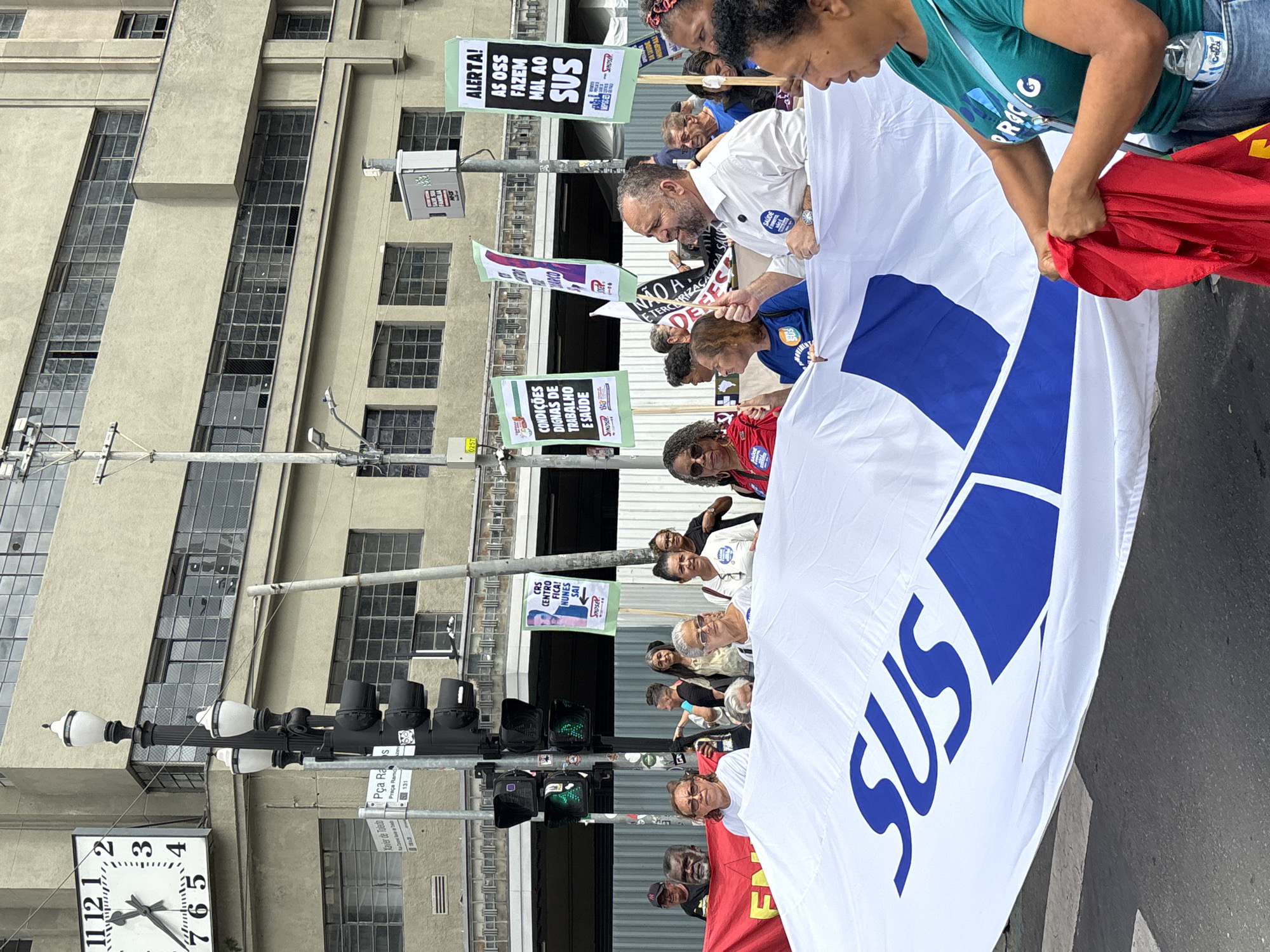 The image shows a street protest in São Paulo, Brazil. Participants hold a large white banner with "SUS" in blue, referring to the Brazilian public healthcare system. Several people hold signs with political slogans. The image also features elements of the urban environment such as a building with multiple windows, streetlights, traffic lights, and a clock showing the time. One of the signs bears the inscription "CRS Centro Fica! Nunes Sai!". Another sign says "Condições Dignas de Trabalho e Saúde." A sign reading "Xavier de Toledo Pca Ra Praça Ramos 131" indicates the location.
