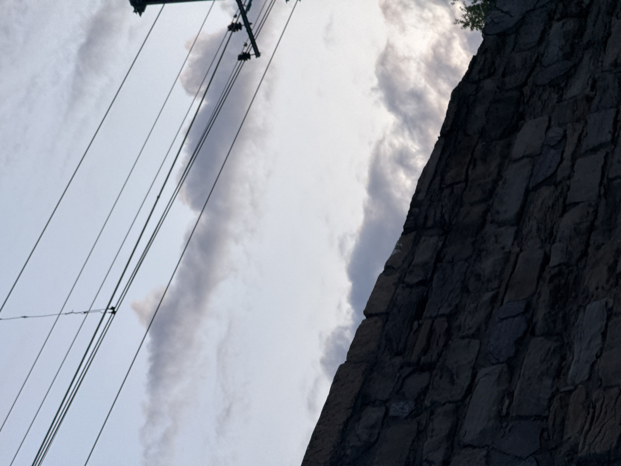 The image shows a partial view of a stone wall on the right side, utility wires and a pole on the left, and a cloudy sky in the background. The wall appears to be part of an older structure. The utility wires are strung along a pole with insulators. The photo is taken at an upward angle. The exact location in Perm, Russia, is not evident from the image alone.