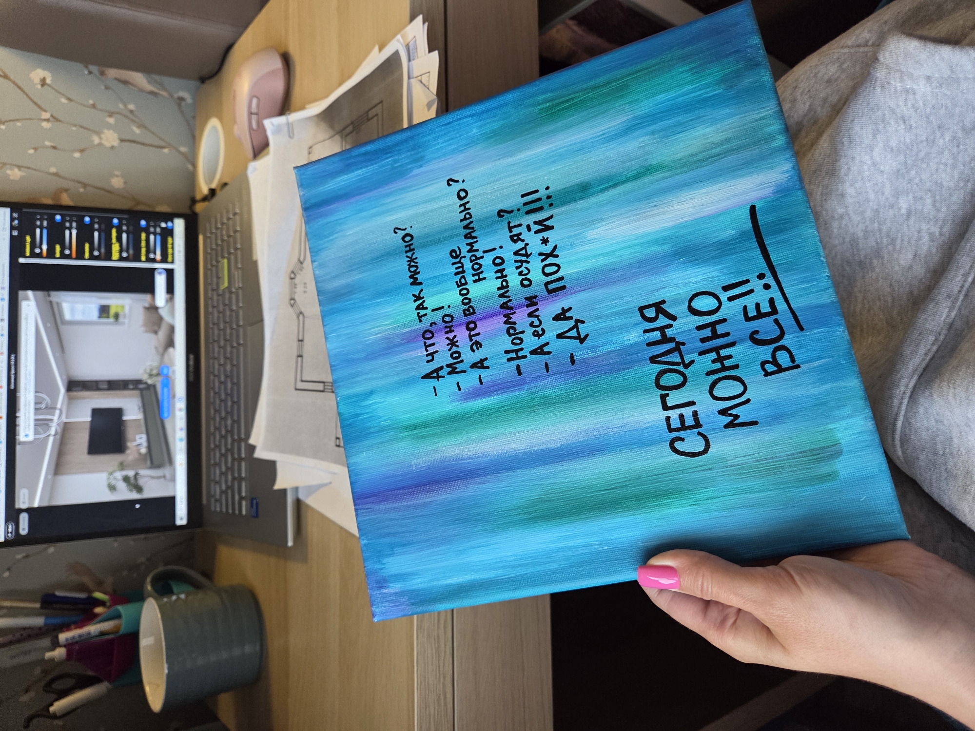 A hand with bright pink nail polish holds a canvas painted with vertical strokes of blue and green. On the canvas, black lettering reads: "А что, так можно? Можно! А это вообще нормально? – Нормально! – А если осудят? – ДА ПОХ*Й!!!" The lower portion of the canvas has larger lettering reading: "СЕГОДНЯ МОЖНО ВСЕ!!". The canvas is positioned over the lap of a person wearing a grey sweatshirt. In the background, a laptop displays an interior design rendering. A grey mug and a pen holder with multiple pens are visible on a wooden desk to the left of the laptop. A pink computer mouse rests to the right of the laptop. Papers, possibly architectural plans, are stacked beneath the canvas and laptop. The scene is likely indoors in a home or office environment. There is no indication this image is located in Birobidzhan, Russia.