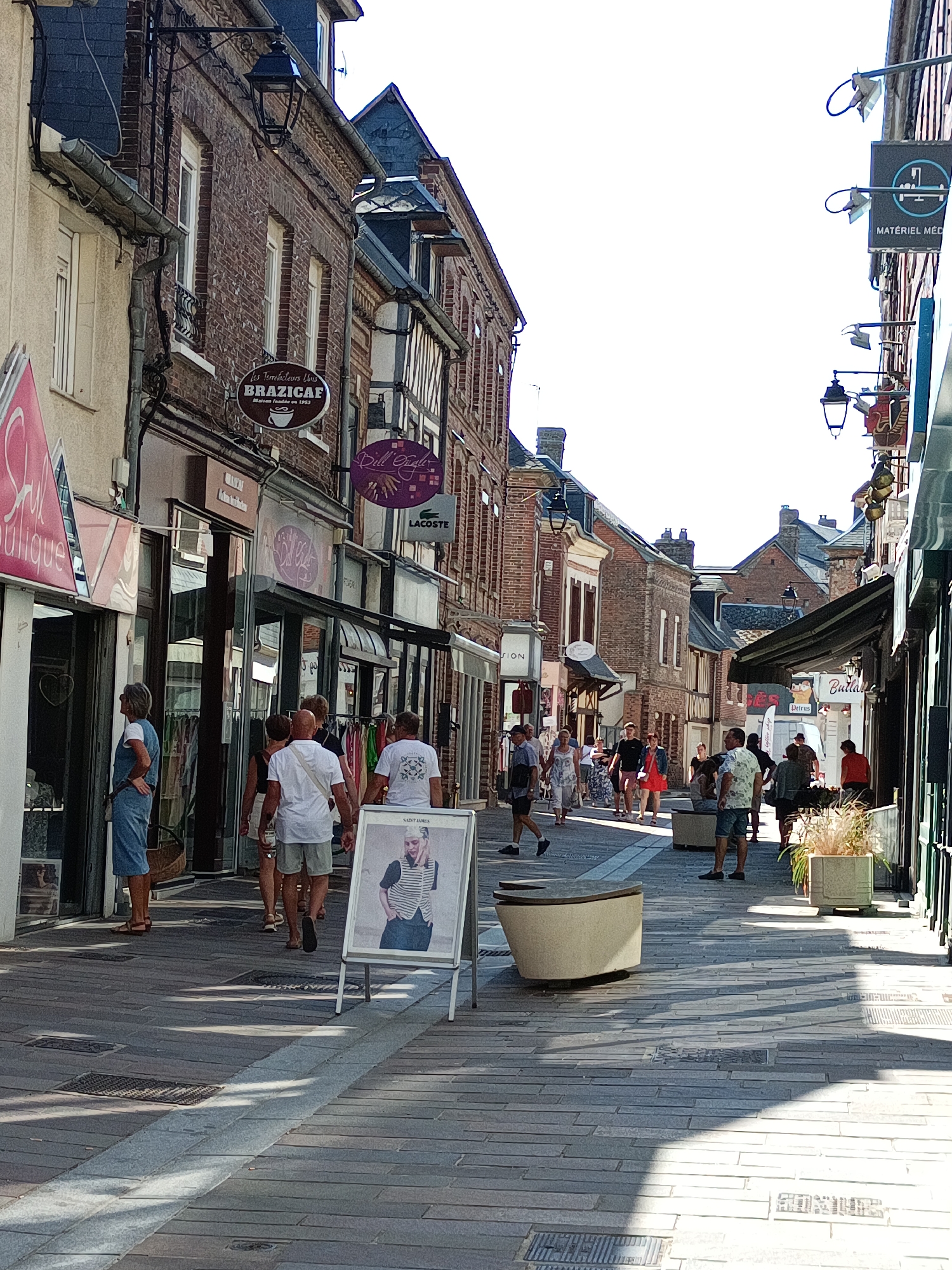 The image depicts a street scene in Le Neubourg, France, likely during daytime. The street is paved with grey stone slabs, with a distinct white line running down the center. On both sides of the street are buildings constructed with red brick, featuring multiple stories, dormer windows, and decorative architectural elements. Several shops are visible with their storefronts open to the street.
In the foreground, a woman in a blue denim dress and sandals stands looking into a shop. To her right, a man in a white t-shirt and grey shorts walks away from the camera. Further down the street, several other individuals are walking, some in groups, others alone. These include a man in a white t-shirt and shorts, a woman in a floral dress, a couple in casual attire, and several other pedestrians.
Notable signs and shops identified include "Brazicaf Maison fondée en 1953," "Styque Boutique," and "Lacoste." There are also street lamps attached to the buildings, providing illumination. In the middle ground, a white A-frame sign displays a photograph of a person. Two modern, curved concrete benches are placed along the sidewalk. On the right side of the street, a potted plant with tall grass is visible in front of a shop. The overall atmosphere suggests a busy commercial street in a town.