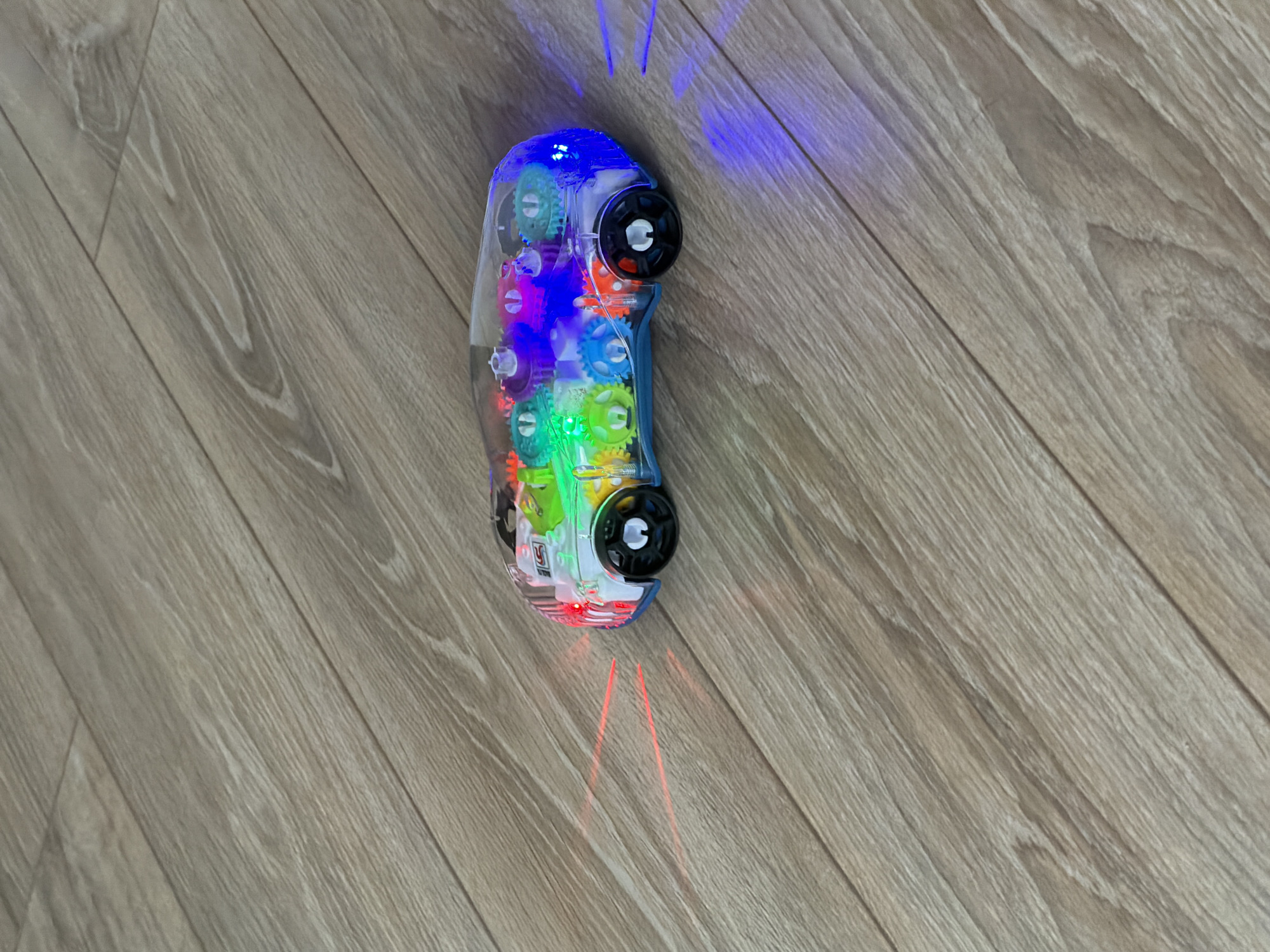 A translucent toy car with illuminated internal mechanisms rests on a light-colored wooden floor. The car's clear plastic casing reveals an intricate arrangement of brightly colored plastic gears in shades of green, blue, orange, pink, and yellow. It has black wheels, two of which are visible on the front and rear.
The toy emits vibrant lights: a bright blue light emanates from its front-top section, casting visible streaks onto the floor, while red light beams project from its rear-bottom, creating distinct lines on the surface. These lights highlight the car's functional or decorative features.
The floor consists of light brown wooden planks with noticeable grain patterns, laid diagonally across the frame. The image is captured from a top-down perspective, emphasizing the car's details and its light projections on the floor. The scene suggests an indoor setting, likely a home. There are no people visible. An indistinct white label with unreadable text is visible within the car's transparent casing.