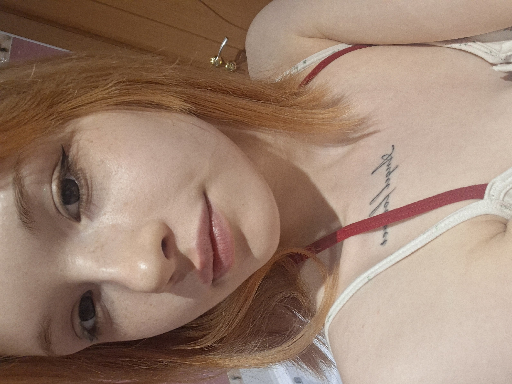 A fair-skinned, red-haired woman with freckles is angled in an indoor space with a wooden background. She wears a white tank top with red straps and has a tattoo on her collarbone. A gold stud earring is visible in her hair. The image does not contain information that would verify a specific location, nor does the environment point to Chaikovsky, Russia.