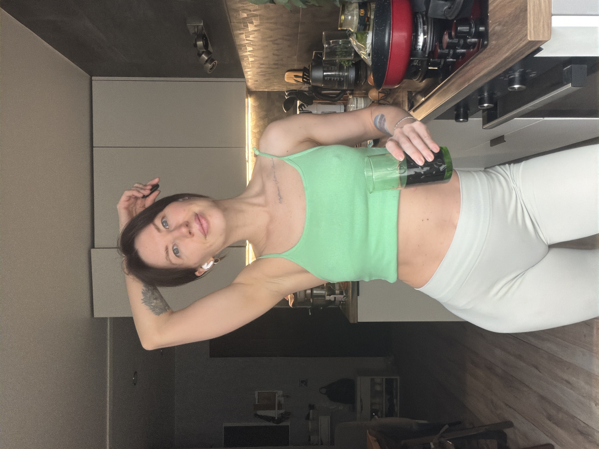 A young woman with short brown hair, wearing a light green tank top and white leggings, is holding a glass filled with a dark liquid in a kitchen. She has a tattoo on her left arm and another on her chest. The kitchen has grey cabinets and a countertop cluttered with appliances and various objects. The location might be a residential apartment in Perm, Russia.