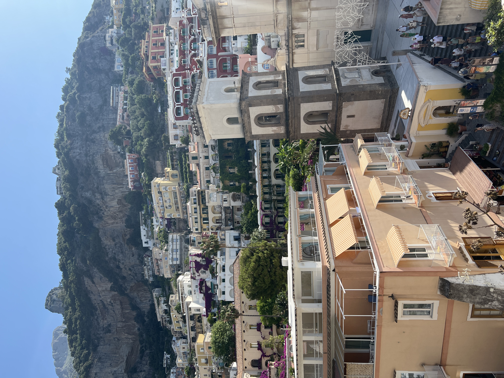 The image depicts a steep, sun-drenched hillside in Positano, Italy, part of the Amalfi Coast. The architecture is characterized by multi-story buildings with pastel facades, white balconies, and colorful awnings, clinging to the rugged terrain. A prominent bell tower, made of stone and stucco, rises from a central piazza, adorned with arched openings. Numerous people, dressed in casual summer attire, are visible walking up and down stone staircases, suggesting a bustling tourist destination. Lush green vegetation and vibrant pink bougainvillea cascade down the buildings and along the cliff face, contrasting with the terracotta rooftops. The overall scene conveys a vibrant and picturesque Mediterranean atmosphere.