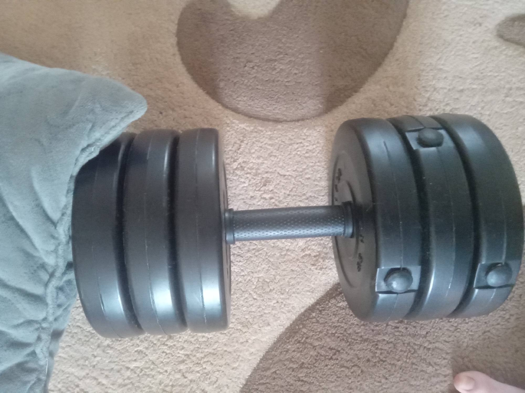A black dumbbell with adjustable weights rests on a textured beige carpet. A light blue blanket is draped over the top of the dumbbell. The dumbbell consists of a central handle with a textured grip, surrounded by stacked weight plates. The weight plates are secured with black clips. A person's thumb is visible in the lower left corner of the image, near the bottom weight plates. The scene appears to be indoors, likely in a domestic setting. No specific location in the city of Terebovlia, Ukraine, can be determined from the image.