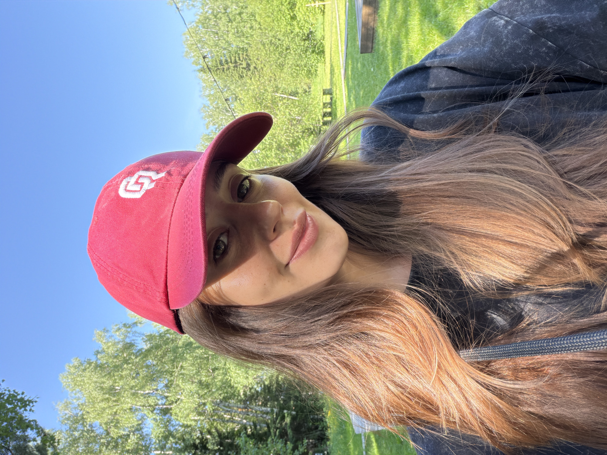 A woman is taking a selfie outdoors on a sunny day. She is wearing a red baseball cap with a white logo and a dark-colored t-shirt. Her long, light brown hair is visible. The background consists of green grass, trees, and a clear blue sky. The image was taken in the city of Minino, Russia.