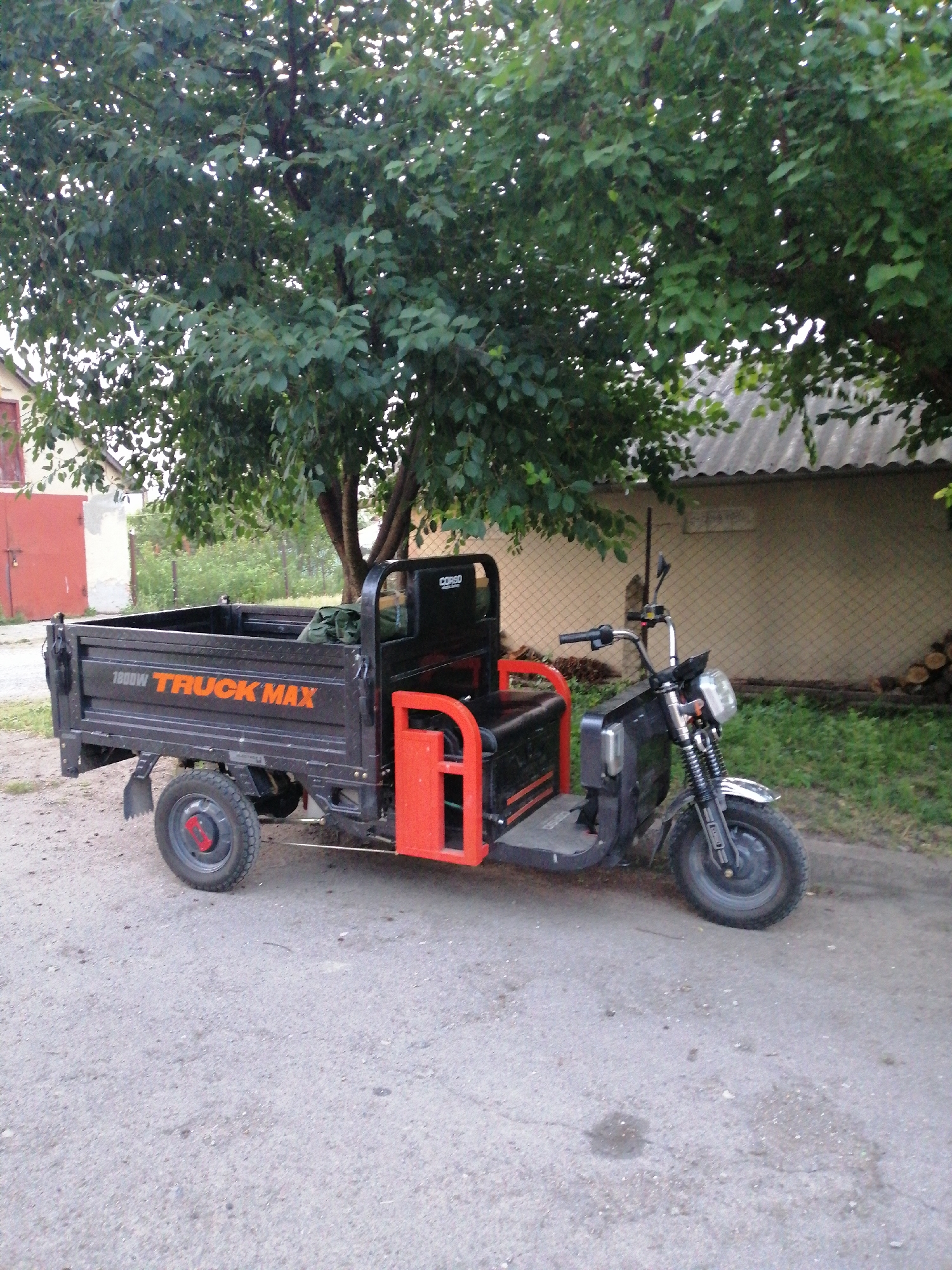 A three-wheeled motorized cargo vehicle, identified by the branding "1800W TRUCK MAX" in orange lettering on its black cargo bed, is parked on an asphalt surface. The vehicle has a black chassis with red accents on the side of the driver's compartment and red metal bars on the rear of the cargo bed. It features a single front wheel and two rear wheels. A green bag is visible in the cargo bed.
The vehicle is positioned in front of a chain-link fence and a building with a corrugated roof. A large tree with dense green foliage shades the scene. In the background, a red metal door and part of a house are visible. To the right of the fence, a pile of chopped wood is stacked. The overall environment suggests a residential or semi-rural setting. The image was taken in the city of Lanivtsi, Ukraine.