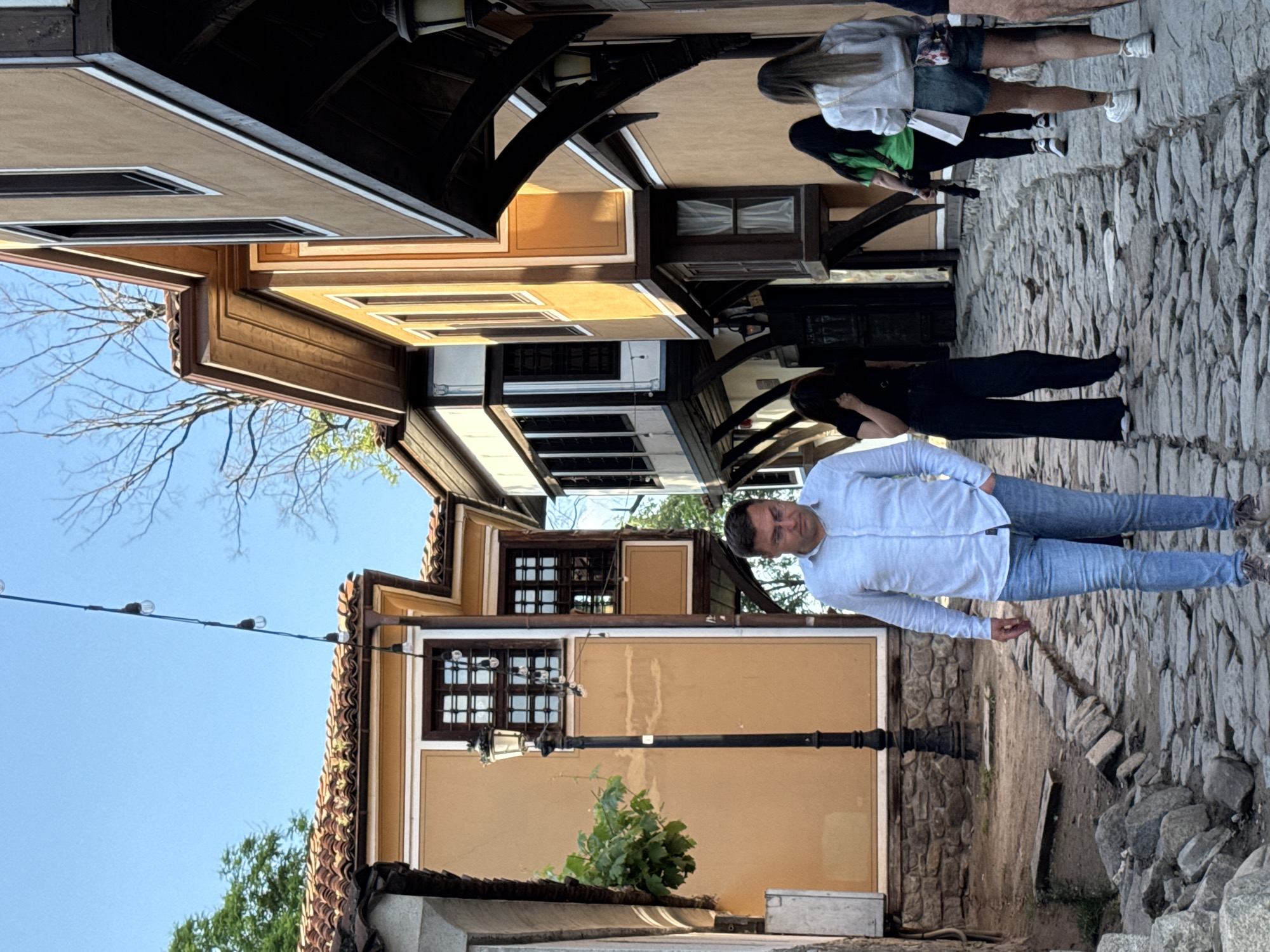 The image depicts a street scene in the old town of Plovdiv, Bulgaria. The street is paved with cobblestones and lined with traditional Bulgarian houses, some with overhanging eaves and wooden architectural details. A man in a light blue shirt and jeans stands in the street, appearing to pose for the photograph. Other pedestrians are visible further up the street, including a woman in a light-colored top and denim shorts, and another person in a green shirt. A lamppost stands near the center of the frame, with vegetation at its base. An overhead wire with bulbs runs along the left side of the image. The sky is clear and blue.