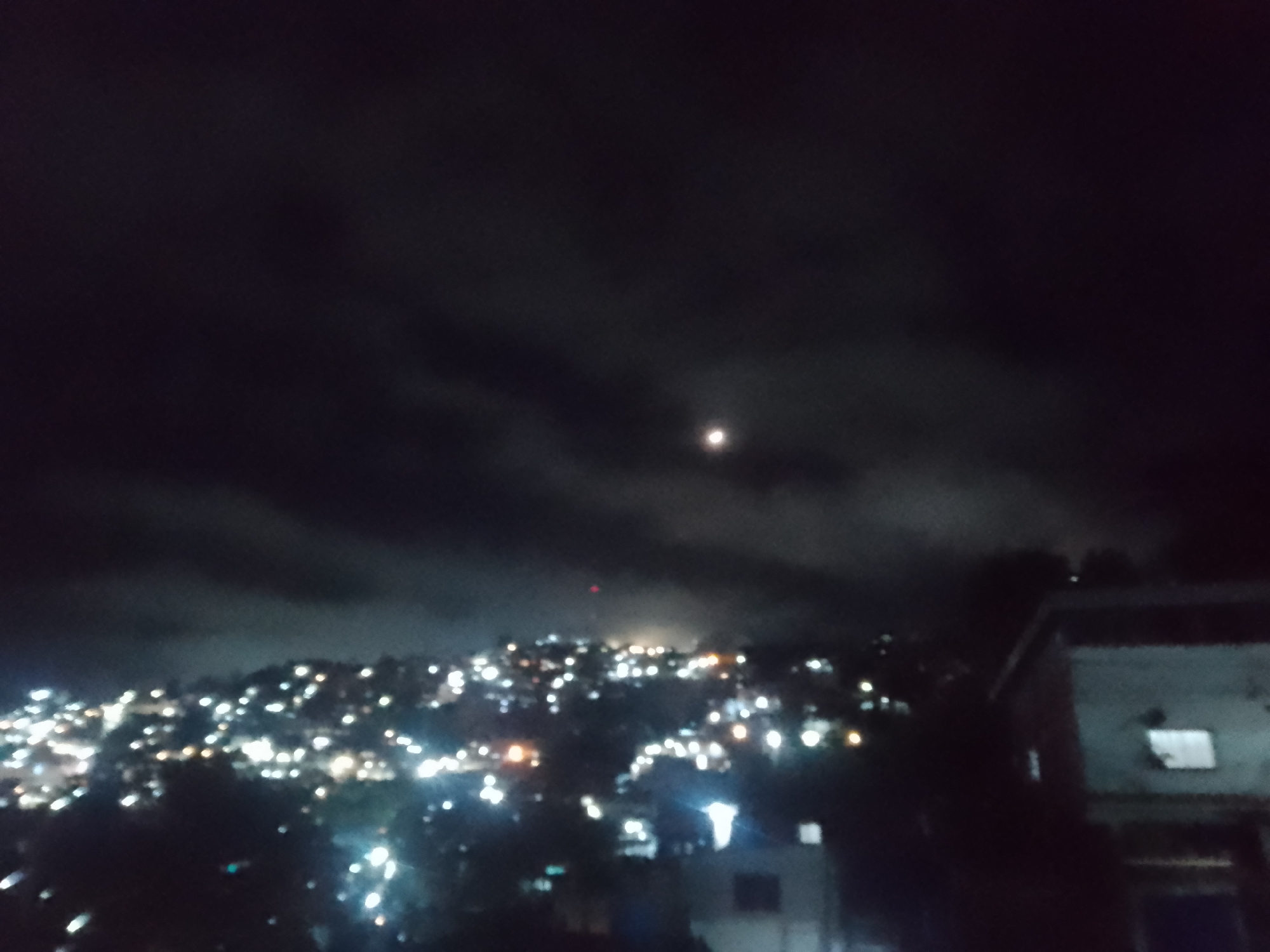 The image depicts a nighttime view of a densely populated hillside in Caracas, Venezuela, illuminated by numerous artificial lights. The sky above is dark and overcast, with a faint moon visible through the clouds. The cityscape sprawls across the terrain, with clusters of lights indicating buildings and streets on the slopes. In the foreground to the right, a portion of a building is visible, its facade dimly lit. The overall impression is of a vibrant, albeit blurry, urban landscape at night, characterized by the pervasive glow of city lights against a dark backdrop.