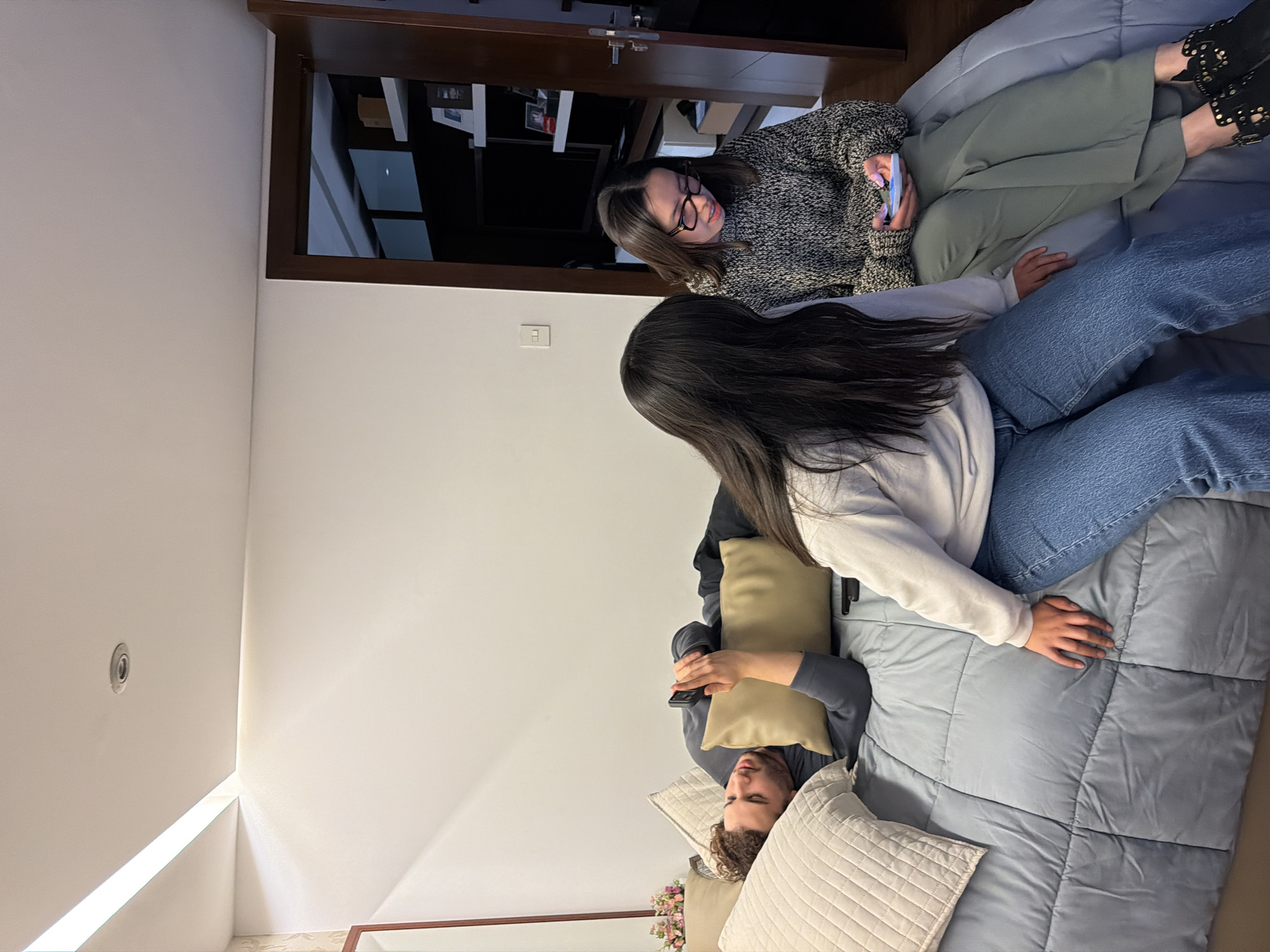 The scene is an interior room in San Bernardino Tlaxcalancingo, Mexico, with three individuals present. A male, identified by curly brown hair and a light complexion, lies on his back on a light blue quilted comforter, wearing a dark grey long-sleeved shirt and dark pants. He holds a dark smartphone in his right hand, resting it on a light brown rectangular cushion. His head is supported by a cream-colored quilted pillow. Seated in front of the male, facing away from the camera, is a female with long dark hair, wearing a light-colored long-sleeved top and blue jeans. To her right, a second female is seated, facing left, wearing dark-framed glasses, a grey and white speckled knitted sweater, and olive green wide-leg pants. She holds a light-colored smartphone with both hands, looking down at its screen. Her feet are clad in black studded ankle boots. The room features white walls, a ceiling with a recessed linear light strip and a circular downlight, and a dark wooden door frame on the right leading to an adjacent room with a dark shelving unit, various objects, and a large dark screen. A partial view of a mirror with a dark frame and a small floral arrangement is visible on the far left. A white electrical outlet or switch panel is mounted on the right wall. The individuals are in close proximity but display no direct physical or verbal interaction, each appearing to be independently engaged with a mobile device or in repose.