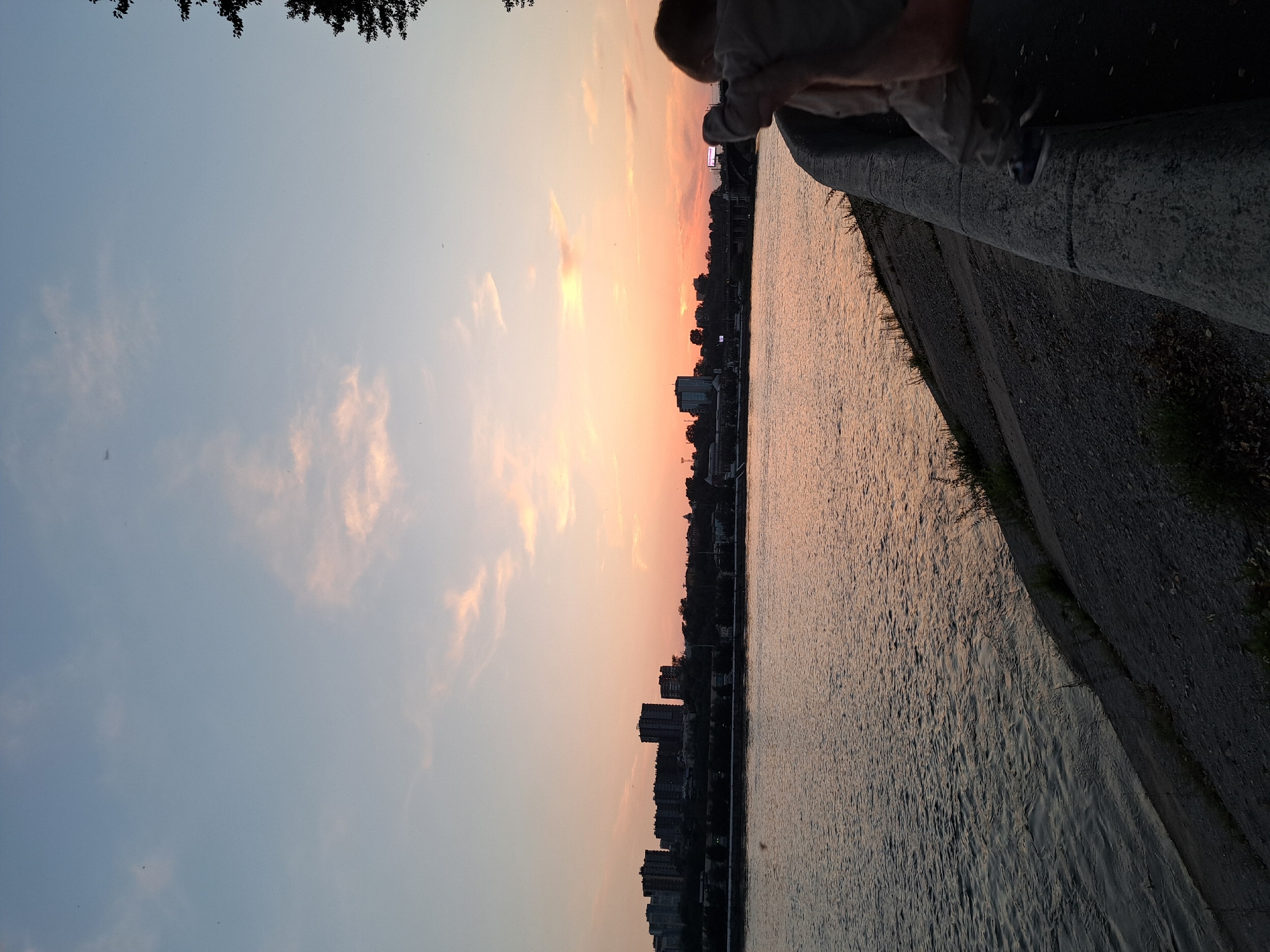 This is a landscape photograph taken at sunset. The image depicts a wide river in the foreground, reflecting the warm hues of the setting sun. In the middle ground, a city skyline with numerous buildings is visible across the river. The sky above is a gradient of pale blue to soft orange and pink, with scattered clouds. A person, partially visible from behind, is standing on a concrete embankment in the foreground, facing away from the camera and towards the river and city. The overall scene suggests an urban waterfront environment during the evening.