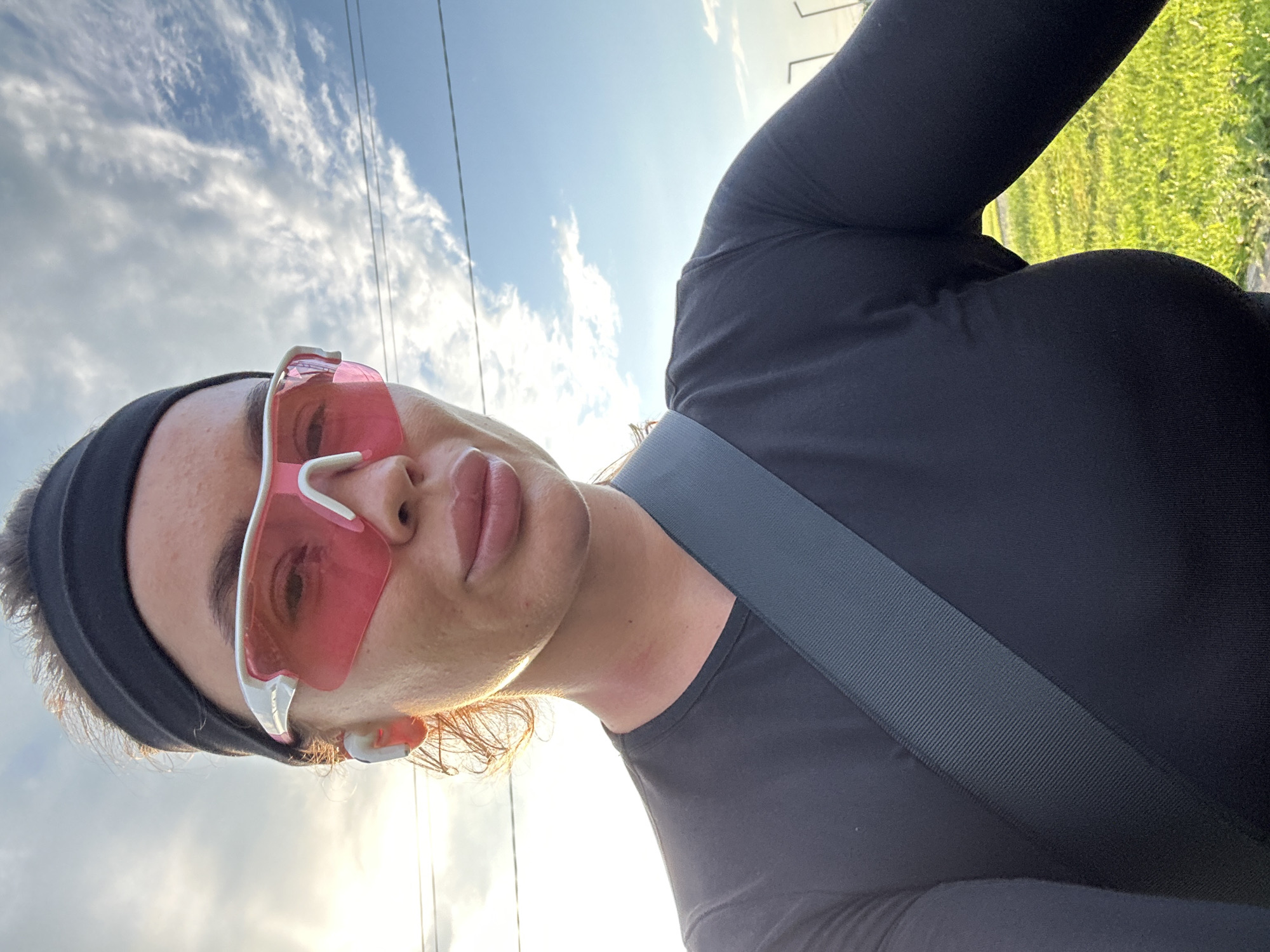 A close-up, low-angle, selfie-style shot shows a person wearing a black long-sleeved shirt and a black headband. They are also wearing large pink-tinted sport sunglasses with white accents. A grey strap crosses diagonally across their chest. The person appears to be outdoors, with a bright, partly cloudy sky visible in the background. Sunlight is shining, creating highlights on the person's face and clothing. The sky suggests it is daytime, possibly late afternoon or early evening due to the warm light. Green grass is visible in the upper right corner of the image, indicating an outdoor environment like a park or field. No visible text is present in the image.
