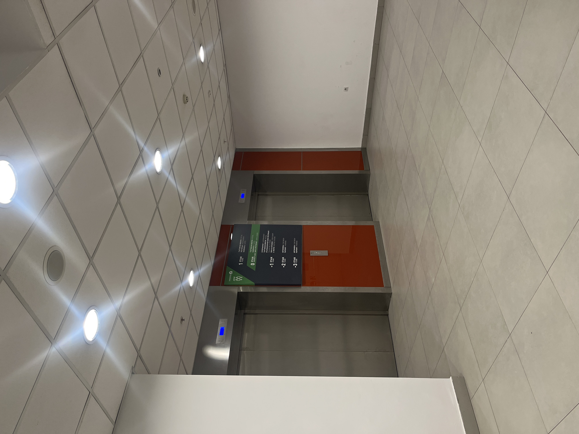 The image shows an interior space with two elevators. The elevator doors are stainless steel, with an orange panel above each. A directory sign is located between the elevators, displaying floor designations in both English and Bulgarian. The first elevator (top) has a blue indicator above it. The ceiling is tiled with recessed lights, and the floor is covered in large, light-colored tiles. The overall environment suggests a modern building. The location is in the city of Sofia, Bulgaria.