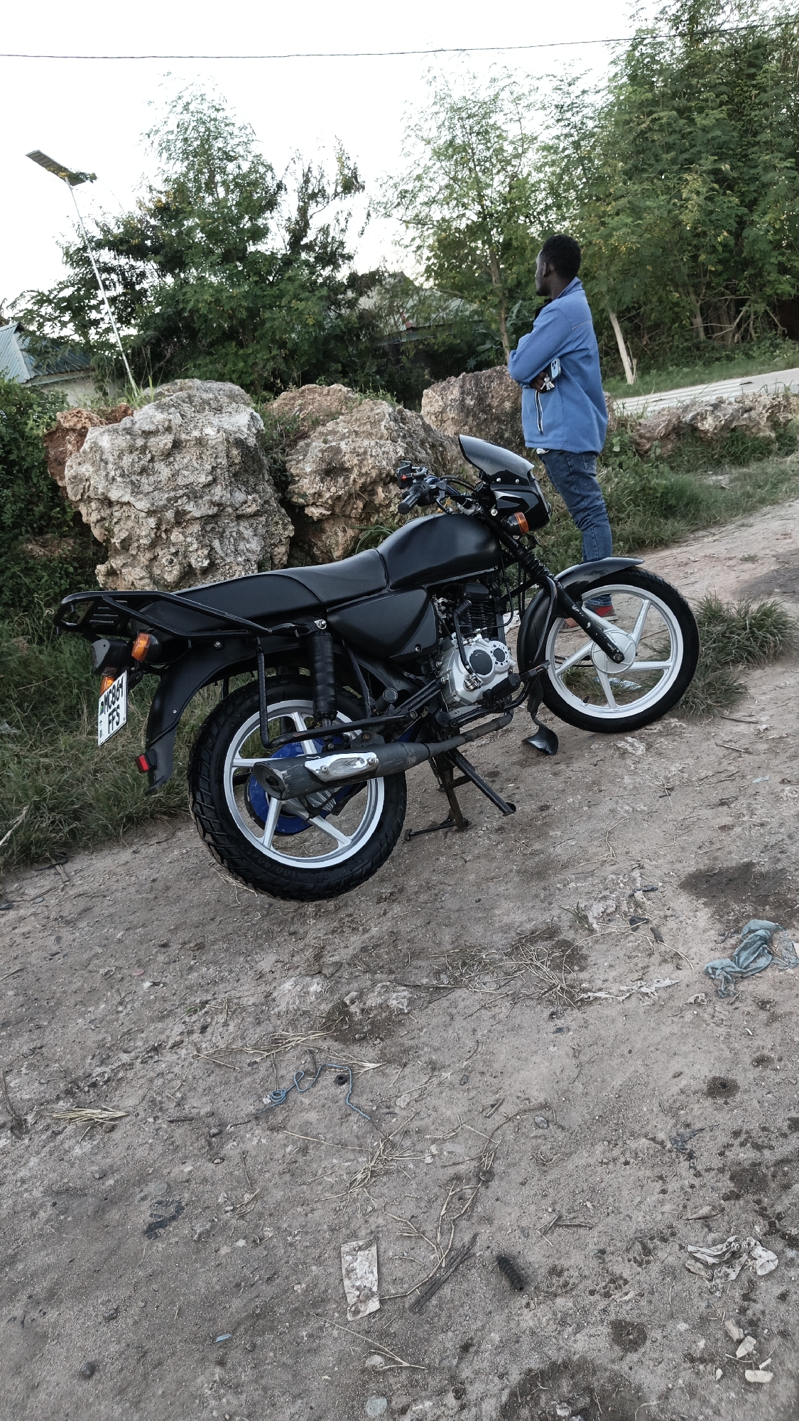 A black motorcycle is parked on a dirt road in Dar es Salaam, Tanzania. A man stands beside the motorcycle, with his arms crossed and looking off into the distance. Behind the motorcycle and the man are large rocks and greenery. In the background, there is a building with a lamppost next to it. Debris is scattered on the ground.
