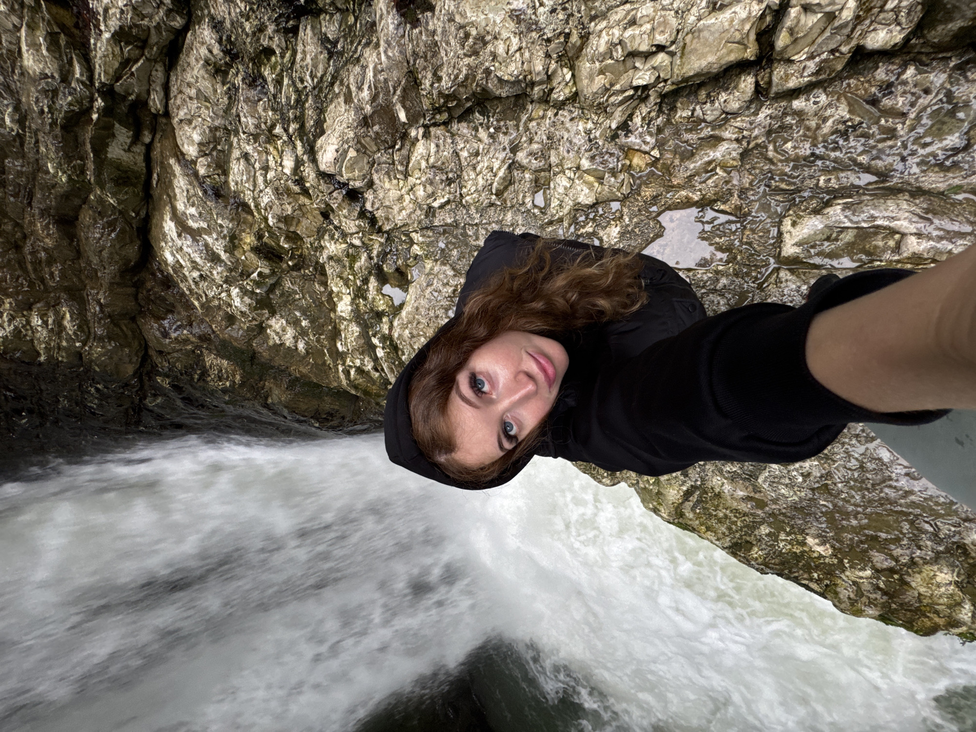 A woman in a black hooded jacket is holding a camera and taking a selfie while standing near a waterfall. The location appears to be a natural environment with rocky cliffs and fast-moving water. The image provides a top-down perspective, showcasing the woman's face and the surrounding landscape, including the waterfall and the rocky terrain. The context suggests this is likely at a natural location, potentially a scenic overlook. Based on the prompt, it's presumed the location is near the city of Tsabal, Abkhazia.