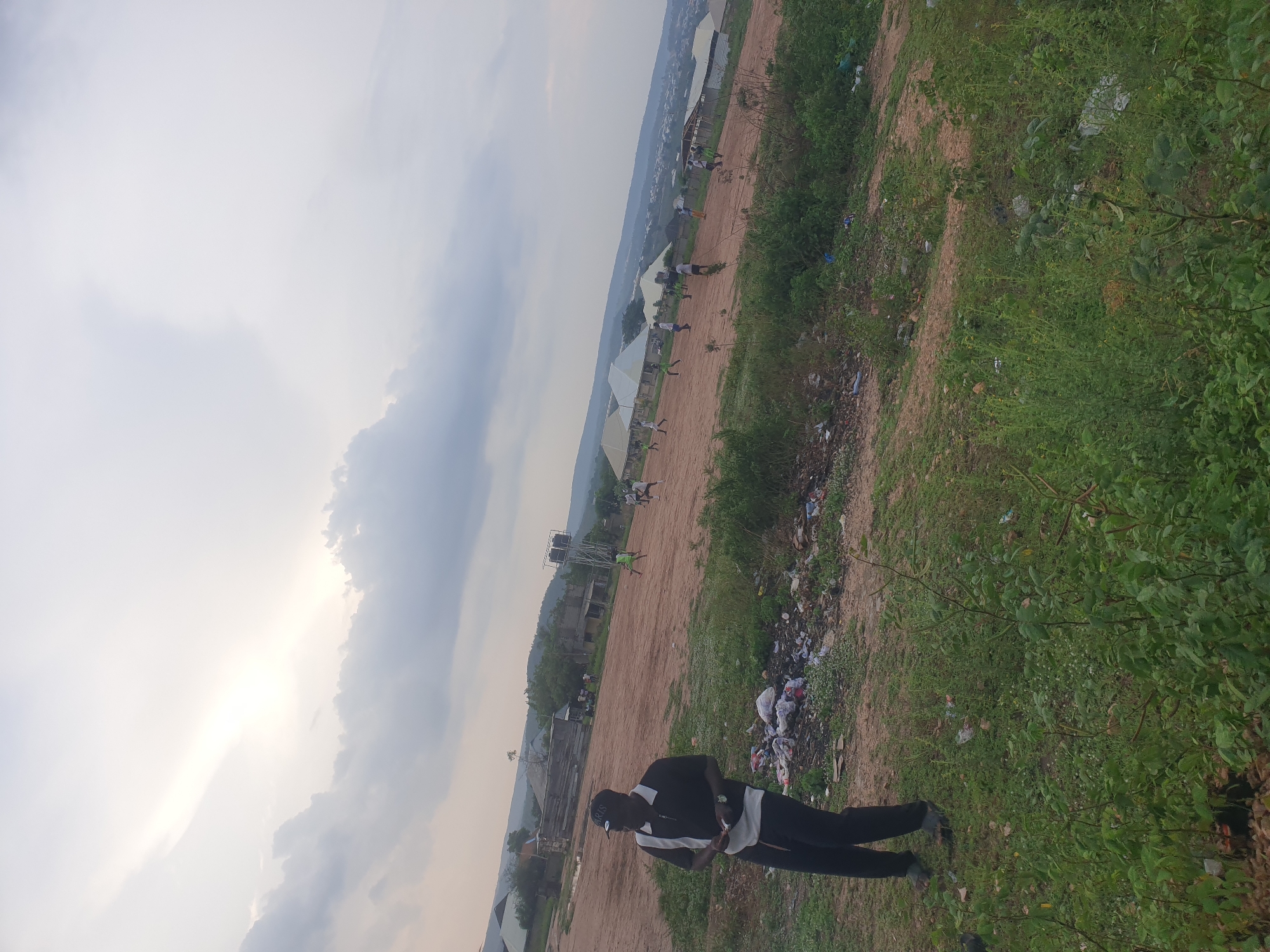 A man is standing on a dirt path. He wears dark pants and a black and white top. There is trash on the path. A line of tents extends along the path into the distance. Other people are further down the path. Buildings and hills are in the background. The sky is overcast. The location is Zango-ndaji, Nigeria.