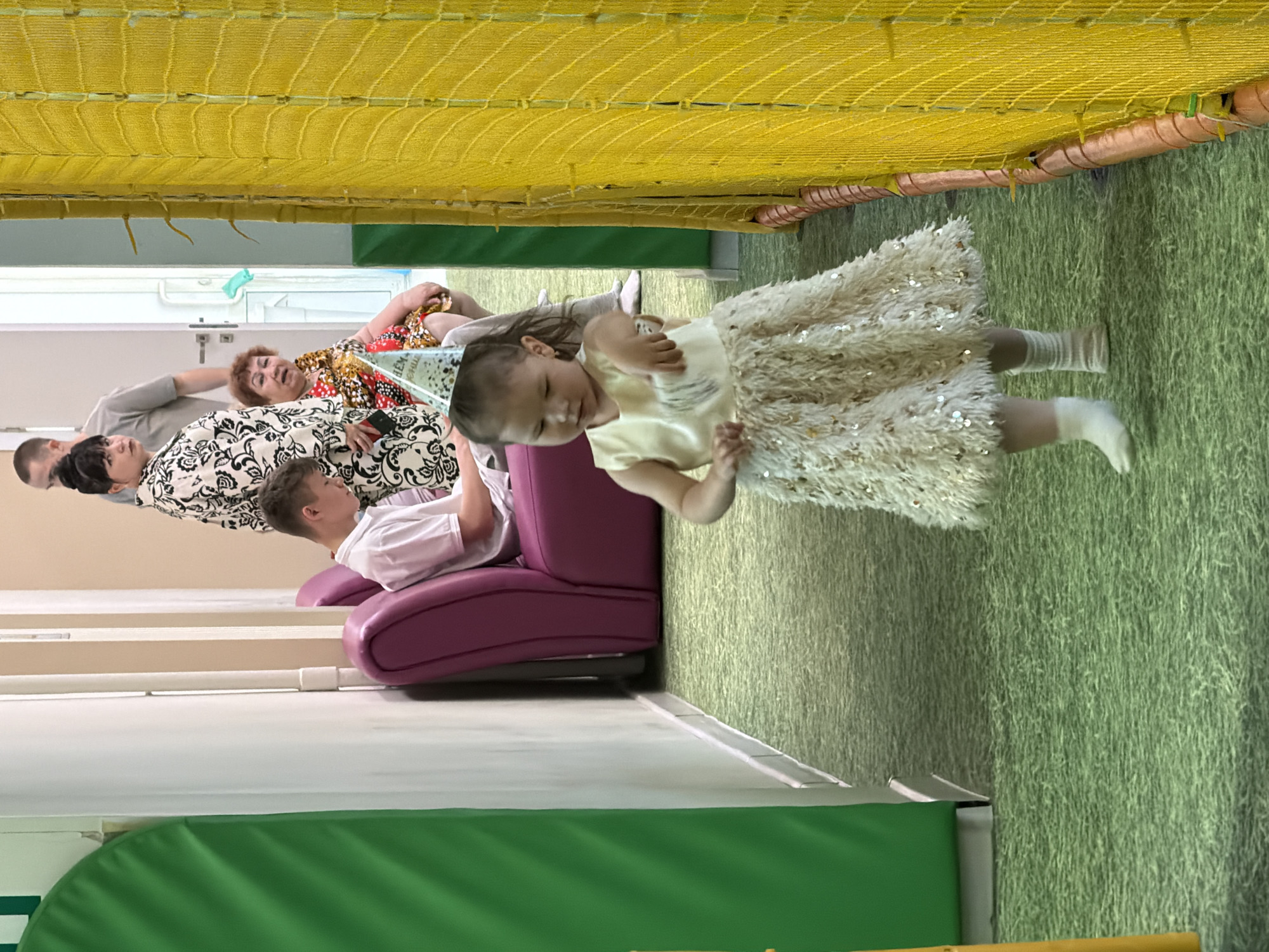 In an indoor play area with green carpeting and walls, a young girl wearing a festive party hat and a white sequined dress is in the foreground, facing right and appearing to dance. Behind her, a teenage boy sits on a purple couch, looking down at his phone. Further back, a woman in a black and white patterned outfit and an older woman in a red and gold patterned dress are standing. The background features a bright yellow net enclosure and a green padded wall. The location is the "Sun City" indoor play center in Chaikovsky, Russia.