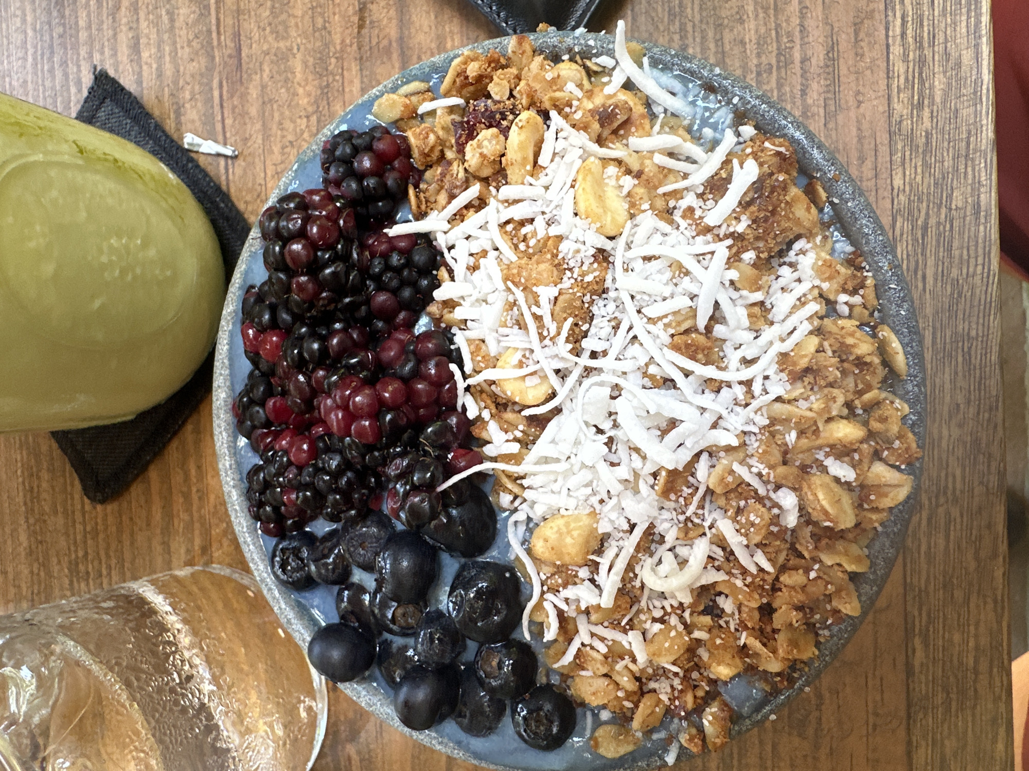 On a wooden surface sits a blue-gray bowl containing a meal of mixed berries, granola, and shredded coconut. The berries include blackberries and blueberries, divided within the bowl. To the left of the bowl is a greenish-yellow liquid in a glass container resting on a black napkin. Part of a glass jar is also visible in the lower left corner of the image. The setting is likely a food establishment in Puerto Escondido, Mexico.