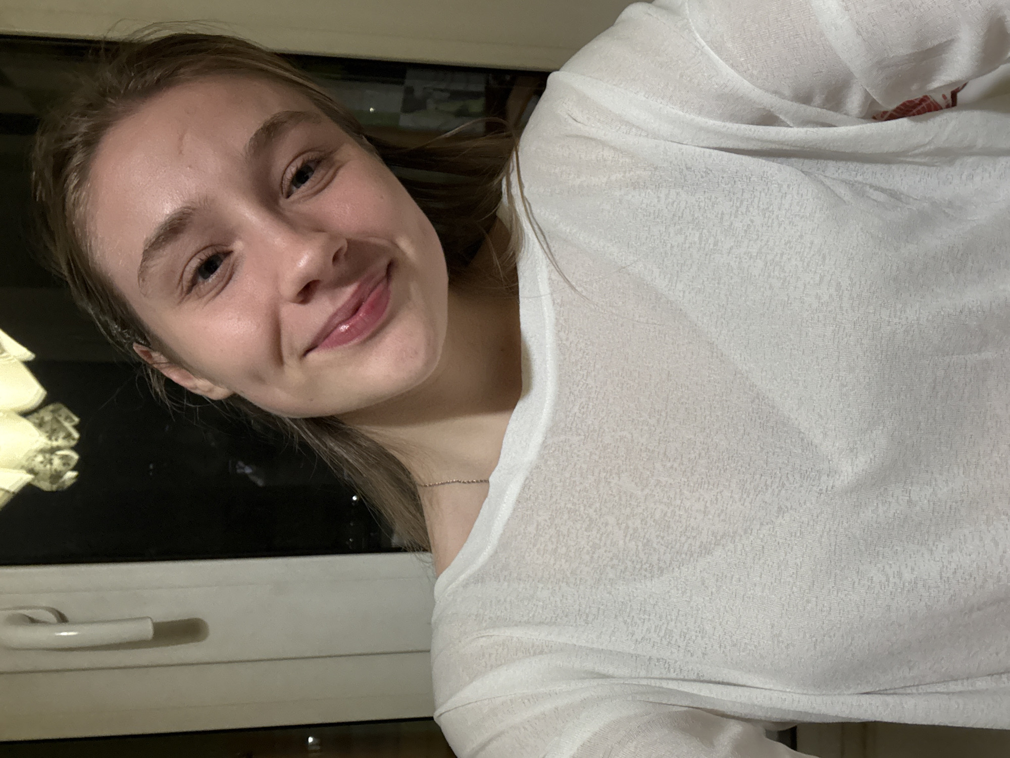 A young woman with fair skin, light brown hair, and a small dimple on her left cheek poses for a selfie. She is wearing a white, semi-transparent shirt. Behind her, a window with a white frame and handle is visible. Outside the window, there are indistinct dark shapes and a light-colored object. The location in the city of Izhevsk, Russia cannot be determined from the image.