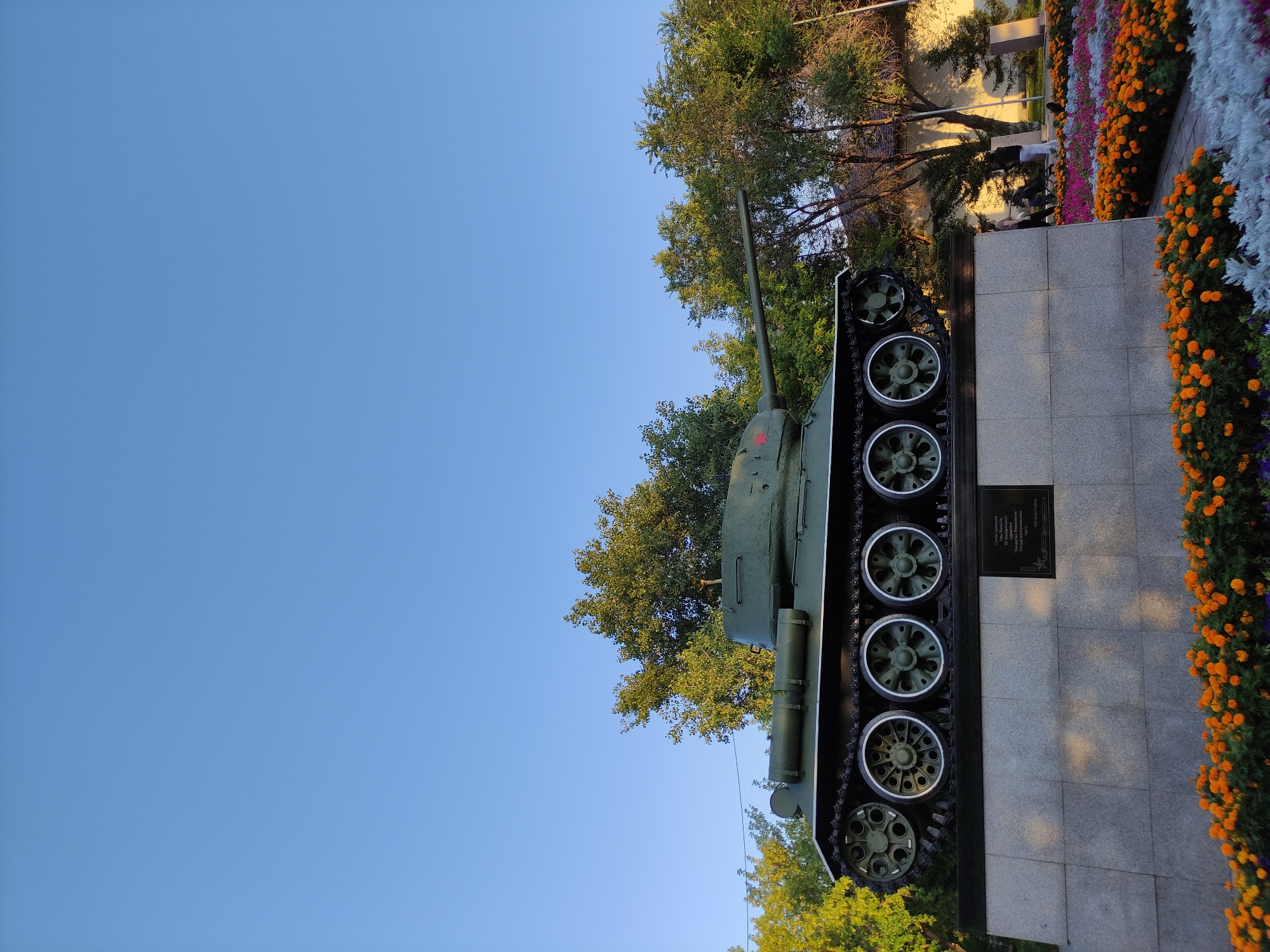 A Soviet-era T-34 tank monument stands on a granite pedestal, set against a clear blue sky. The tank is painted olive green and features a red star emblem on its turret. In front of the pedestal, a vibrant flower bed displays rows of orange, purple, and white flowers. Behind the tank, several large trees with lush green foliage are visible. In the background, a light-colored building with windows and a fence can be seen, along with a few people walking in the distance. The monument is located in the city of Semey, Kazakhstan.