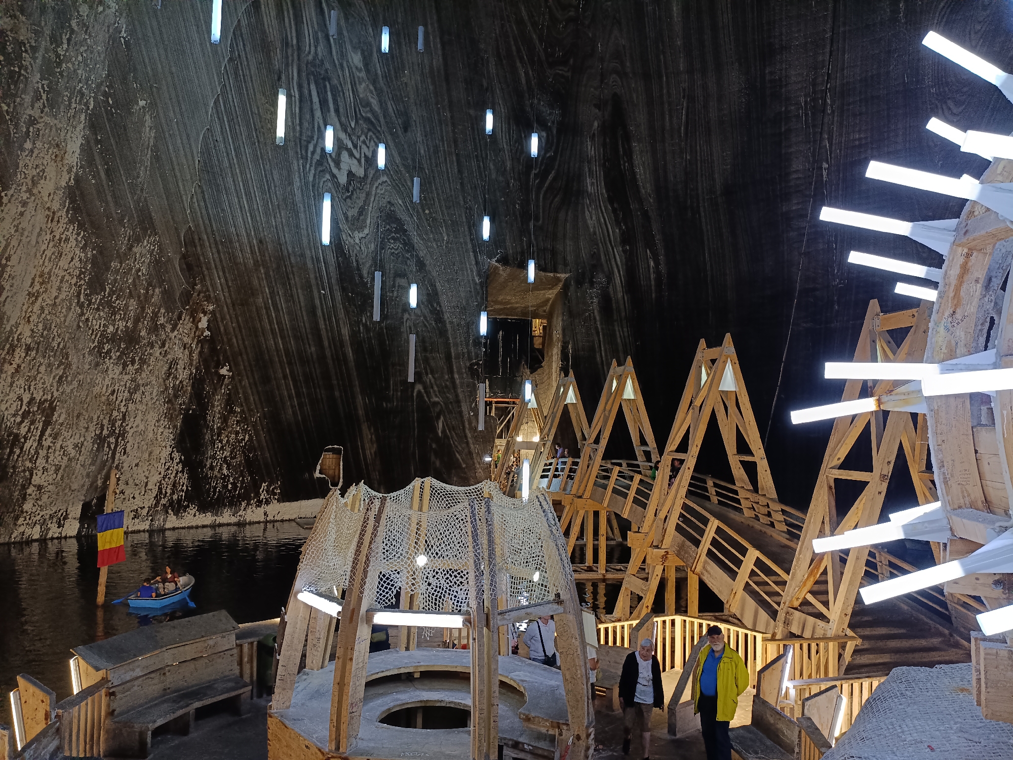 The image depicts the interior of the Salina Turda salt mine in Turda, Romania. The cavernous space is illuminated by numerous hanging cylindrical lights and light strips integrated into wooden structures. In the foreground, a wooden, multi-tiered structure with netting and seating is visible. To the left, a body of dark water is present, with a small blue boat carrying at least two individuals and a Romanian flag on a pole. Further into the background, complex wooden walkways and elevated platforms, also adorned with lighting, ascend the cavern walls. Several people are observed walking on these platforms and near the foreground structures. The salt mine walls, characterized by their stratified, textured appearance in varying shades of gray and brown, form the backdrop of the scene.