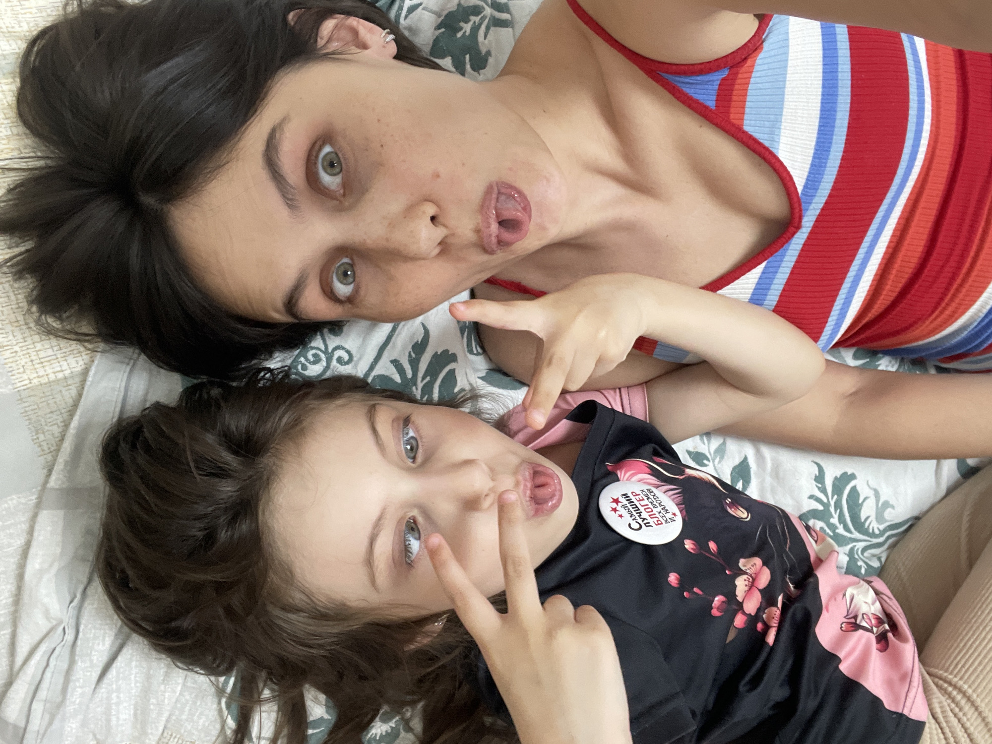 Two females are lying on a patterned bedspread, both making faces with their tongues protruding. The older female has dark hair and wears a striped red, white, and blue tank top. The younger female has brown hair, wears a black t-shirt with pink floral designs, and a button featuring text in Cyrillic. Neither the Izhevsk, Russia city name nor any related landmarks are found within the image.