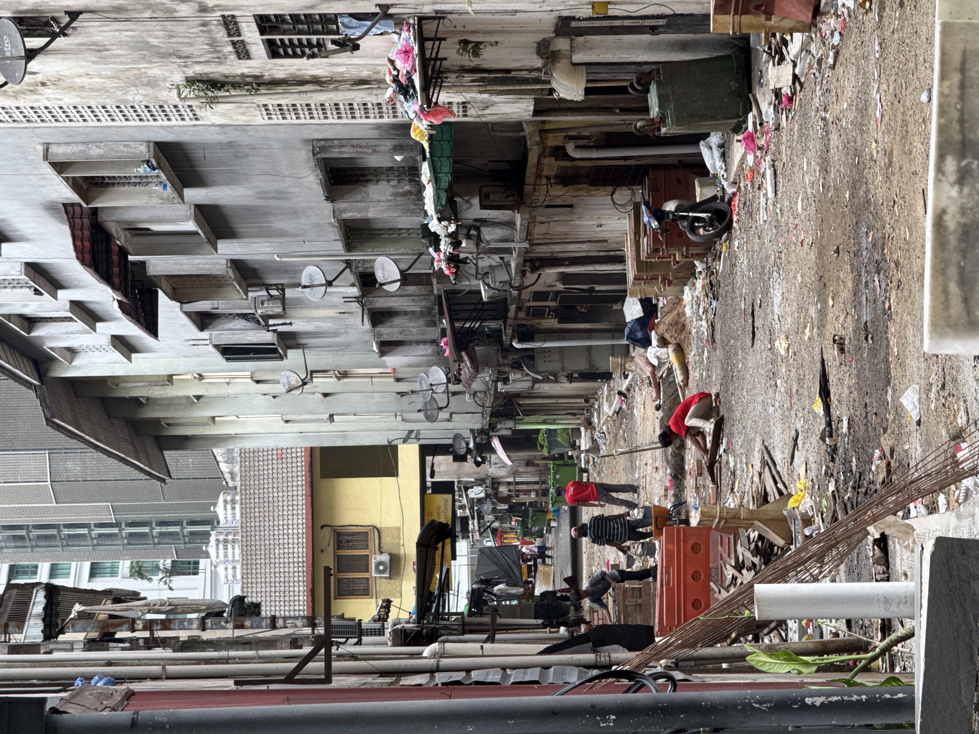 The image depicts a narrow alleyway in Kuala Lumpur, Malaysia, characterized by buildings on both sides, with residents, sanitation workers and garbage on the ground. Overhead, various cables, pipes, and satellite dishes crisscross the alley. The alleyway appears cluttered with discarded materials, trash, debris, and remnants of construction materials. There is a group of workers dressed in casual clothes and PPE equipment along with orange traffic barriers. The buildings lining the alley appear to be aging, showing signs of wear and maintenance, with a mix of architectural styles and functional adaptations. Residents are seen engaging in waste clean-up. The scene suggests a blend of residential and commercial activity, with indications of ongoing maintenance and modifications to the buildings.