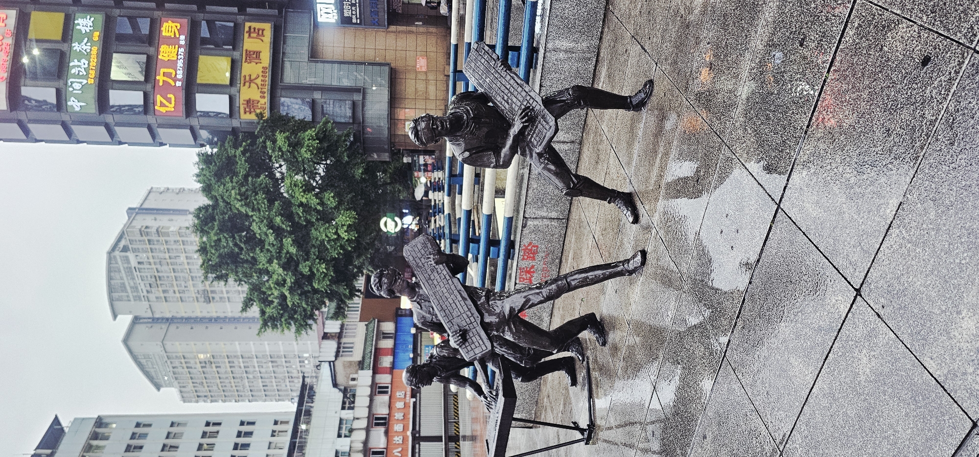 This image captures an urban street scene in Jiulongpo District, China, on an overcast day, characterized by wet, reflective surfaces on the ground, indicating recent precipitation.
In the foreground and midground, three dark, metallic statues of male figures are positioned on a tiled pavement. All figures wear goggles. The leftmost figure stands behind a dark, keyboard-like instrument supported by a metal stand, with its hands positioned as if playing. The middle figure stands, holding a large, elongated object resembling a computer keyboard across its chest, akin to a guitar. The rightmost figure is also standing and holding a similar large, keyboard-like object in a playing posture. The statues' surfaces are glossy and reflect light due to the wet conditions. The pavement consists of large, light gray, speckled tiles, which are uniformly wet and highly reflective, mirroring the sky and surrounding structures.
Behind the statues, a low barrier with alternating blue and white horizontal stripes is visible. Further back, a large, dense green tree stands prominently. To the left of the tree, several buildings rise. Two tall, light-colored modern high-rises with subtly curved rooftops dominate the background skyline, flanked by lower-rise structures.
To the right of the tree, a multi-story commercial building is a prominent feature. Its facade is composed of a grid of square or rectangular panels, many of which display illuminated Chinese characters in various colors. Specifically visible are signs for: "中间站茶楼" (Middle Station Teahouse) in green, "亿力健身" (Yili Fitness) in red and yellow, and "瑞天酒店" (Ruitian Hotel) in yellow, each accompanied by a phone number. A small, circular green and white illuminated sign is visible in the middle distance behind the statues and to the right of the tree. The sky is uniformly light gray and overcast.