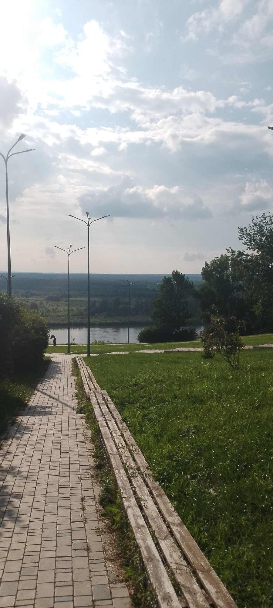 The image depicts a paved walkway in a park setting, likely in Birsk, Russia, bordered by a wooden bench on the right and lush green grass. The walkway, composed of rectangular grey pavers, leads towards a body of water, possibly a river, visible in the mid-ground. Several tall, modern streetlights with multiple lamps are positioned along the walkway and in the distance. Across the water, a dense, dark green forest stretches towards the horizon under a sky filled with scattered white and grey clouds. The sun is partially obscured by clouds, casting a bright light that creates shadows on the walkway. In the far background, a hazy landscape suggests a town or village. On the left side of the walkway, a dark-haired figure, possibly a woman, is standing near a bush, facing away from the camera. The overall scene is tranquil and suggests a public recreational area.
