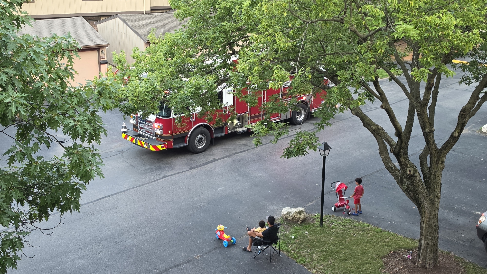 In Elgin, Illinois, a red fire truck is parked on an asphalt road, partially obscured by the lush green foliage of several trees. In the foreground, a man sits in a black folding chair, holding a young child on his lap. Next to them is a red and yellow toy ride-on vehicle. A few feet away, a young boy in a red shirt and shorts stands beside a red stroller. The scene appears to be a residential area, with apartment buildings visible in the background.