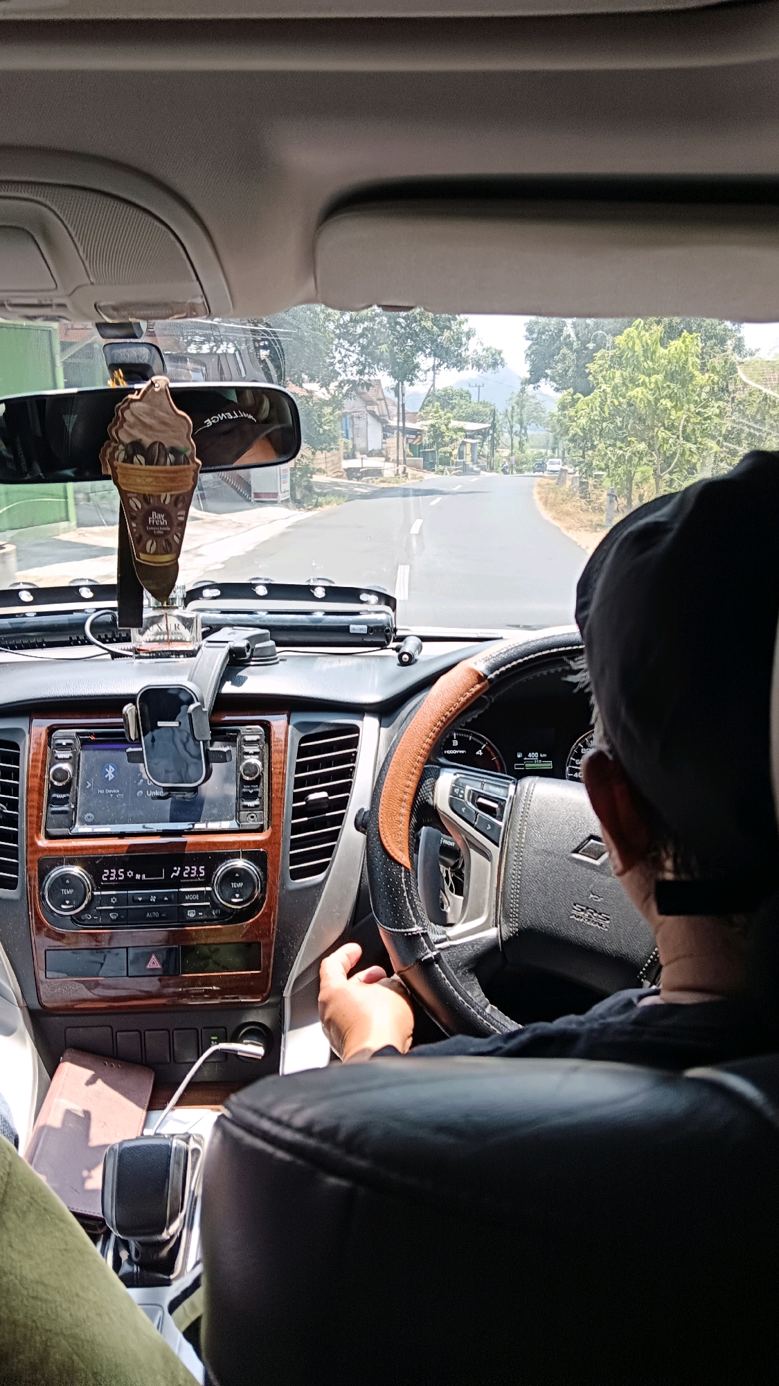 An exterior shot, taken from inside a vehicle, captures a driver behind the wheel on a paved road. The driver, wearing a black cap, is partially visible from the back, with their hands on the steering wheel. The car's dashboard is prominent, featuring a touchscreen display, air vents, and climate controls showing a temperature of 23.5 degrees Celsius. A car air freshener, shaped like an ice cream cone, hangs from the rearview mirror. The road ahead winds through a rural landscape with trees and houses on either side. The scene is set in Kuniran, Indonesia, under a bright, sunny sky.